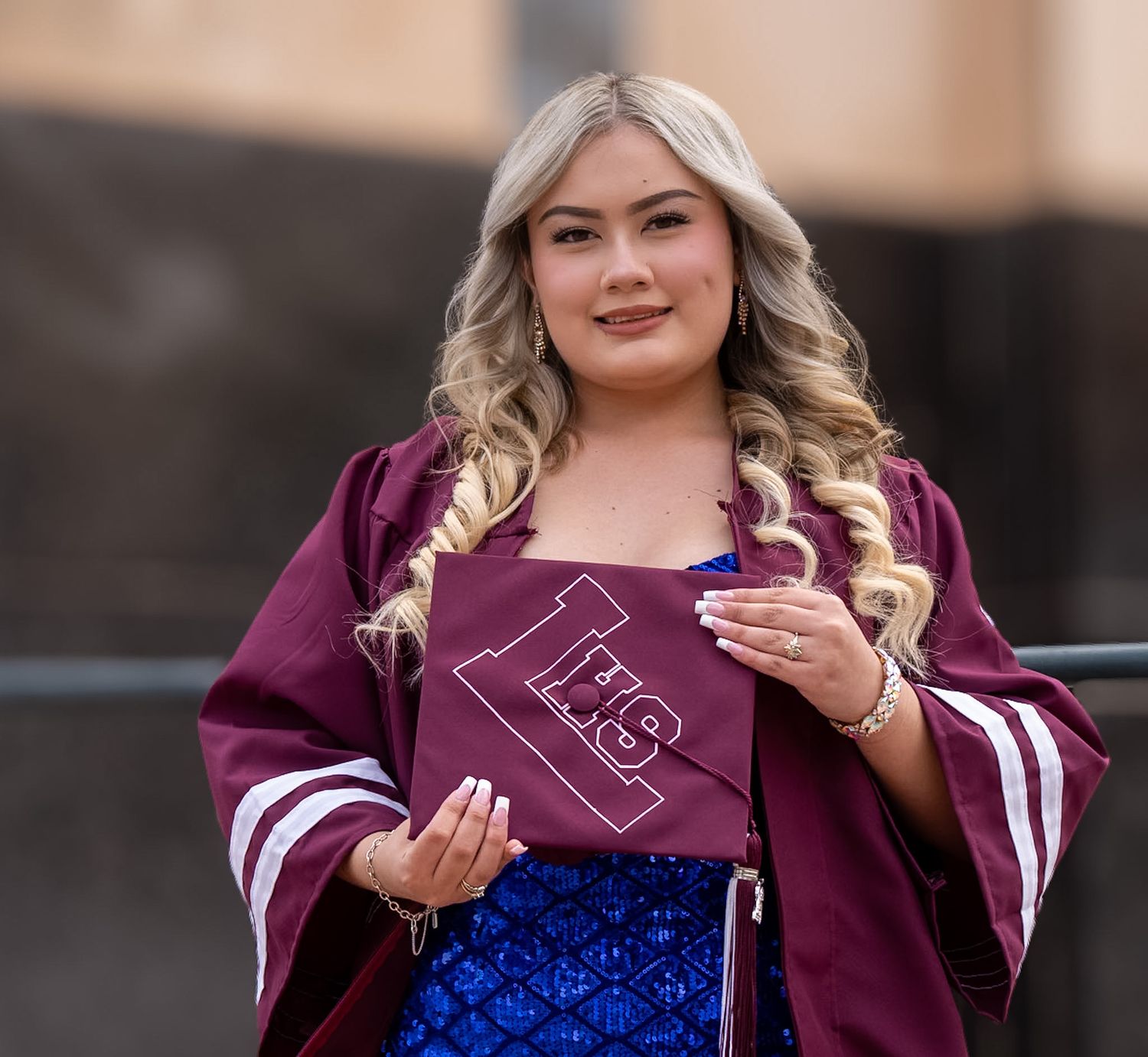 Young woman in maroon graduation gown holding decorated cap, smiling outdoors in blue dress.