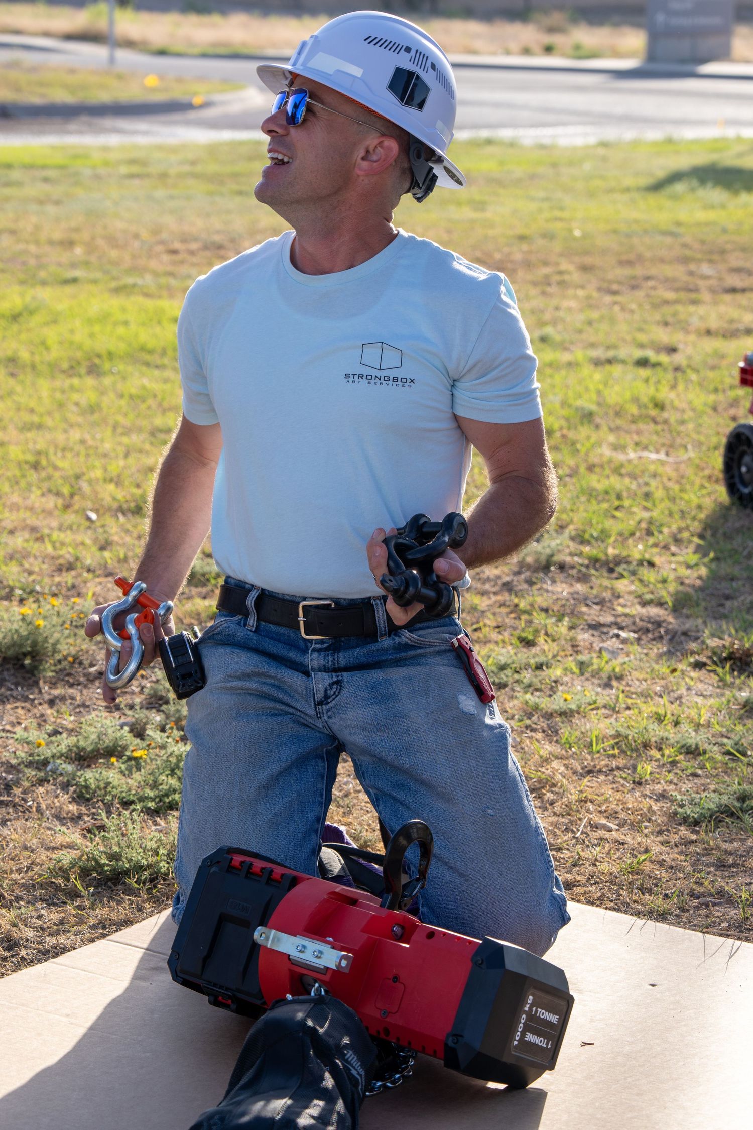 Construction worker in white hard hat kneeling outdoors holding tools near red power equipment.