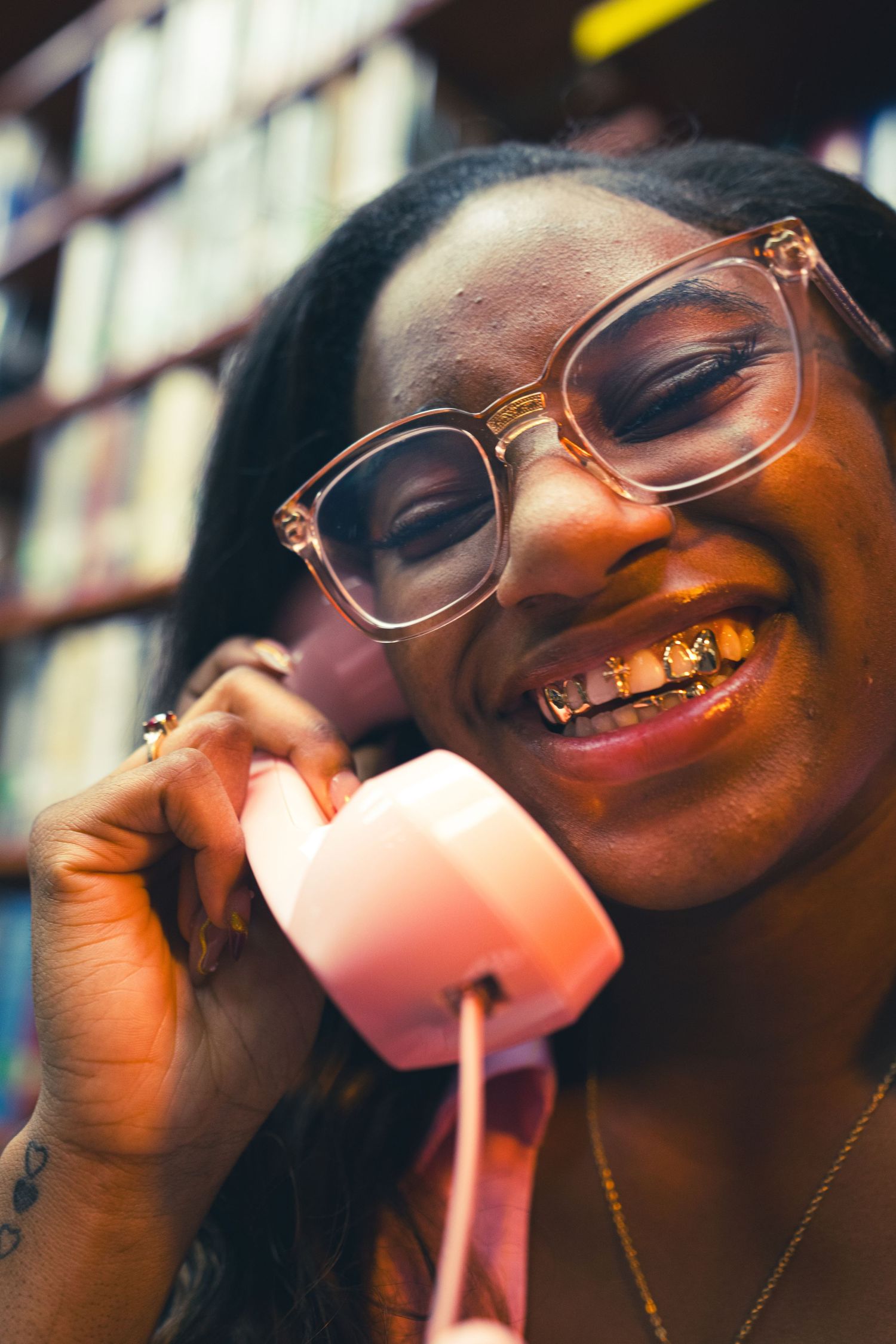 Smiling young woman with braces and clear glasses holds a pink retro telephone handset in a warm, blurred interior.