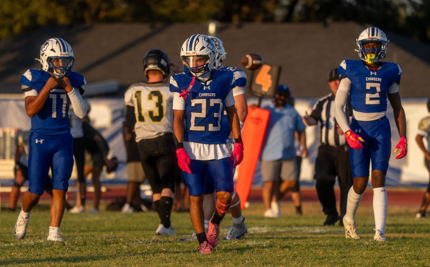 High school football players in blue uniforms with numbers 11, 23, and 2 walk on field during a game.