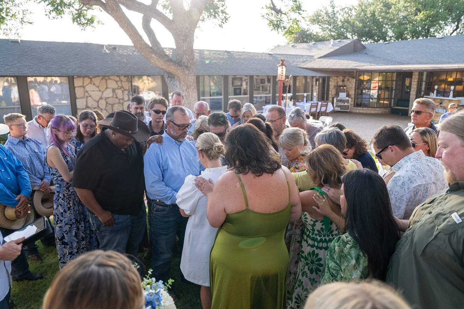 A large group of people gathered outdoors for a community prayer circle near a stone building.
