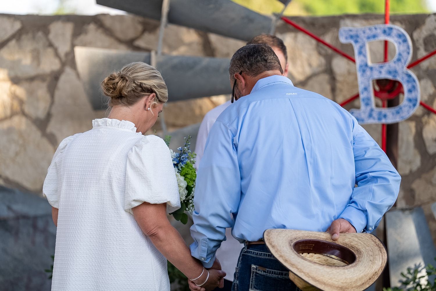 Couple holding hands and bowing heads in prayer near floral arrangement and decorative letter B.