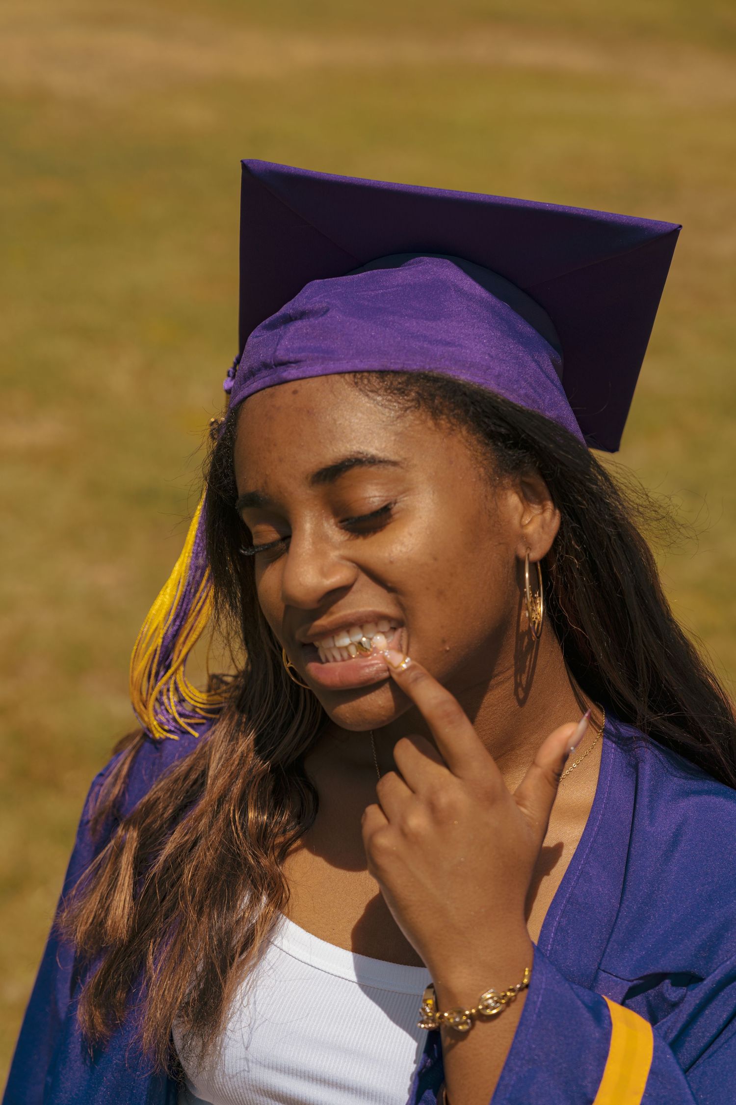 Young Black woman in purple graduation cap and gown smiling with finger on chin outdoors.