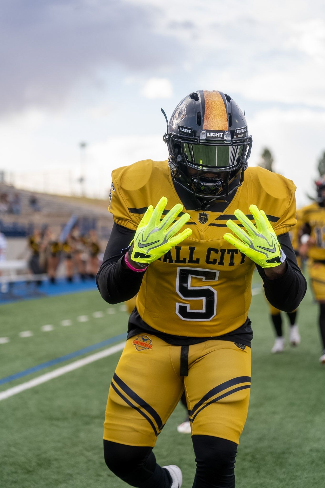 Football player #5 in gold All City uniform poses with hands out under cloudy sky.
