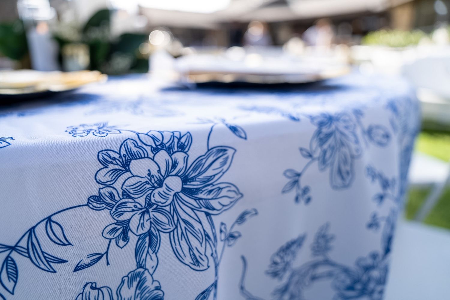 Blue and white floral toile tablecloth draped over an outdoor table set with dark plates in a garden setting.