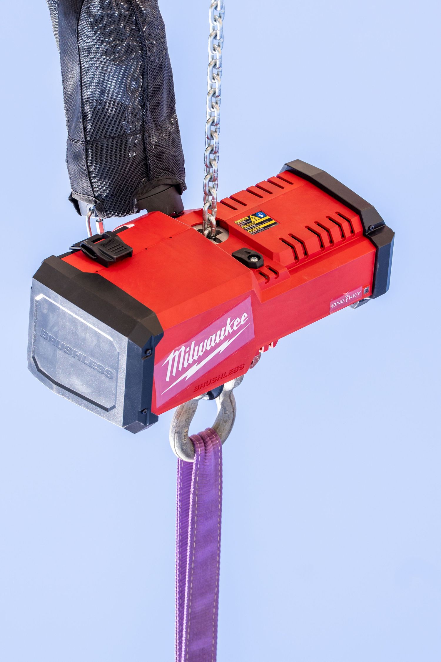 Milwaukee M18 red electric hoist hanging from chain with purple lifting strap against blue sky.