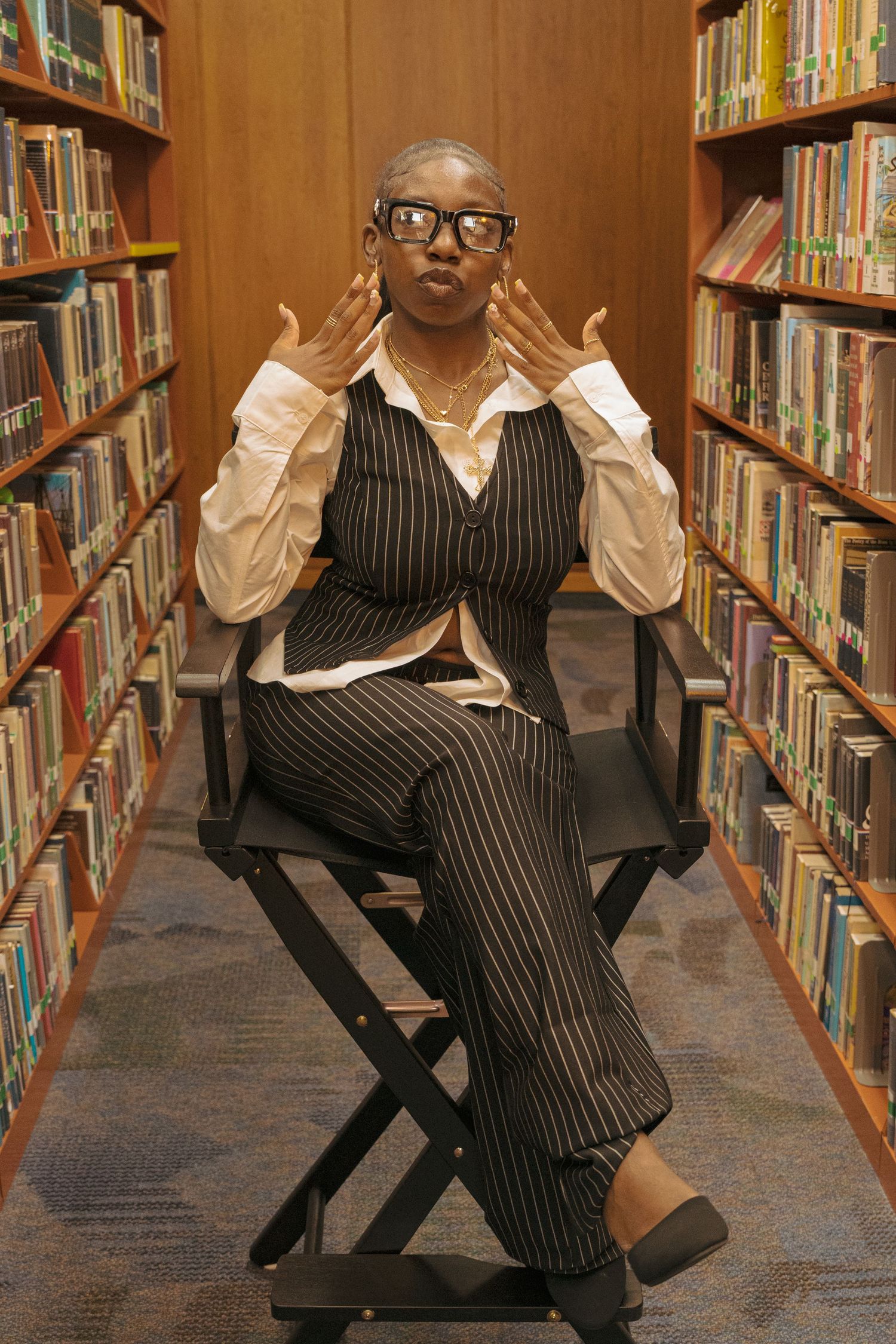 Black woman in pinstripe suit sits in director chair between library shelves making a kissy face.