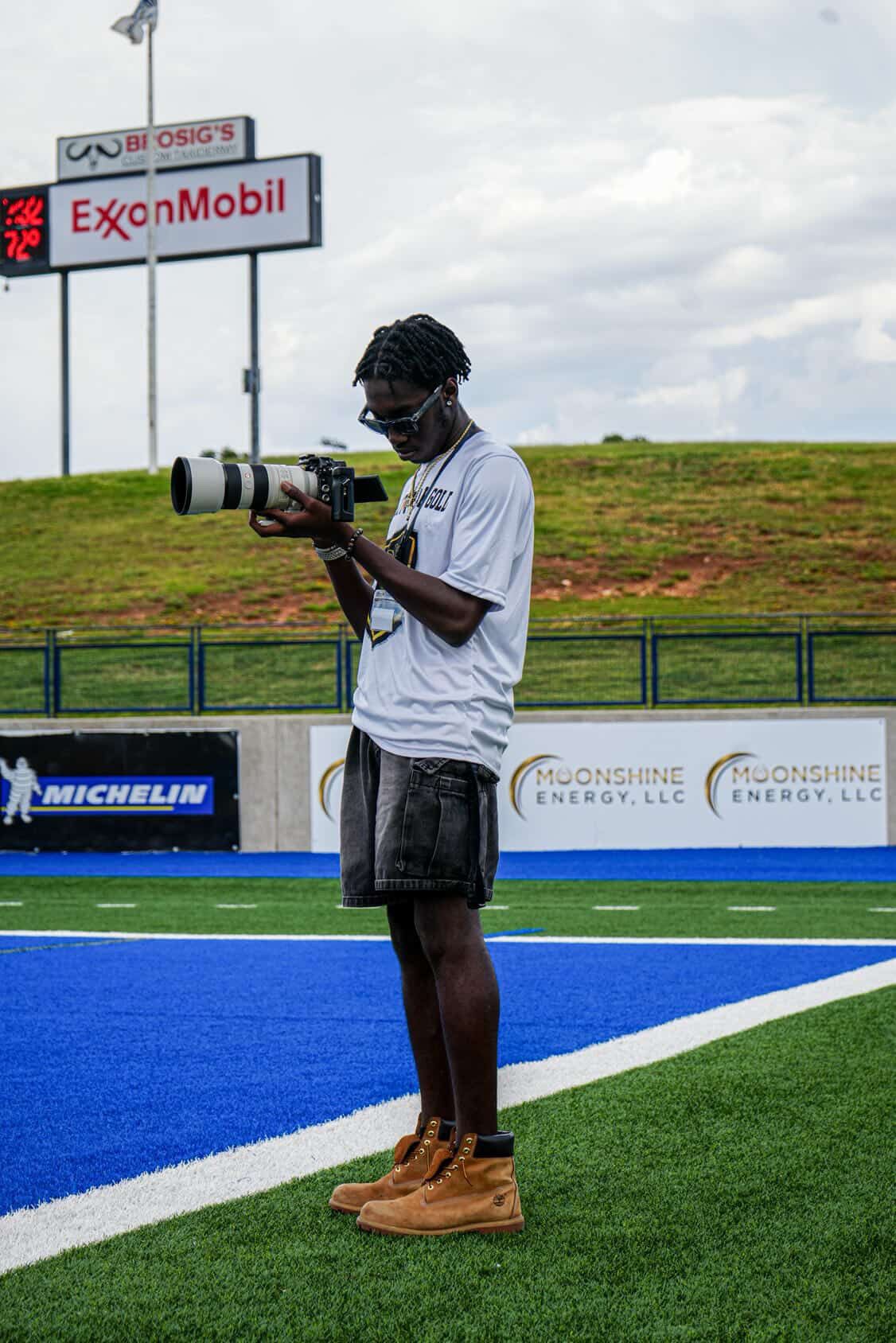 Young photographer reviewing shots on camera while standing on blue turf football field.