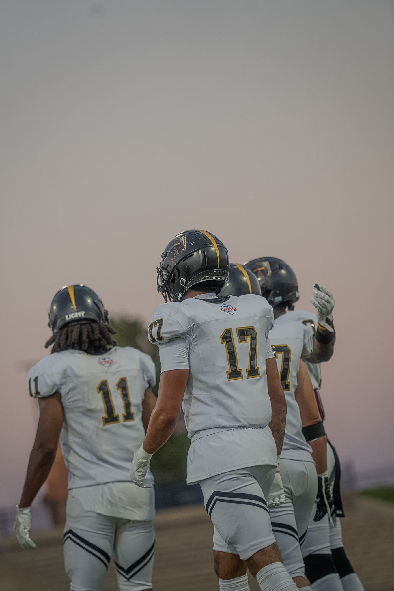 Three Tall City football players in white jerseys numbers 11 and 17 walk at dusk during a game.