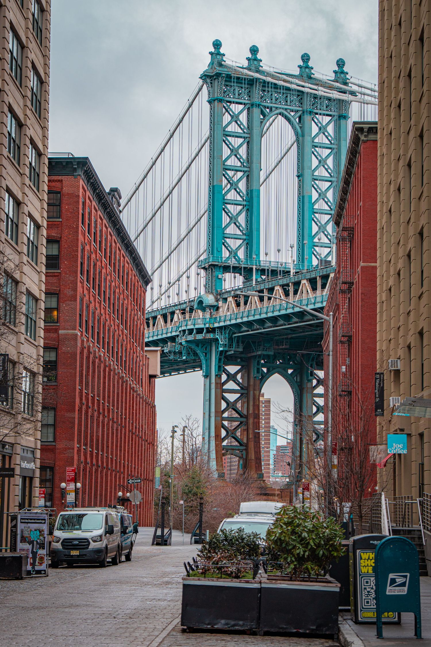 Manhattan Bridge viewed from DUMBO Brooklyn street, framed by red brick buildings on a cloudy day.