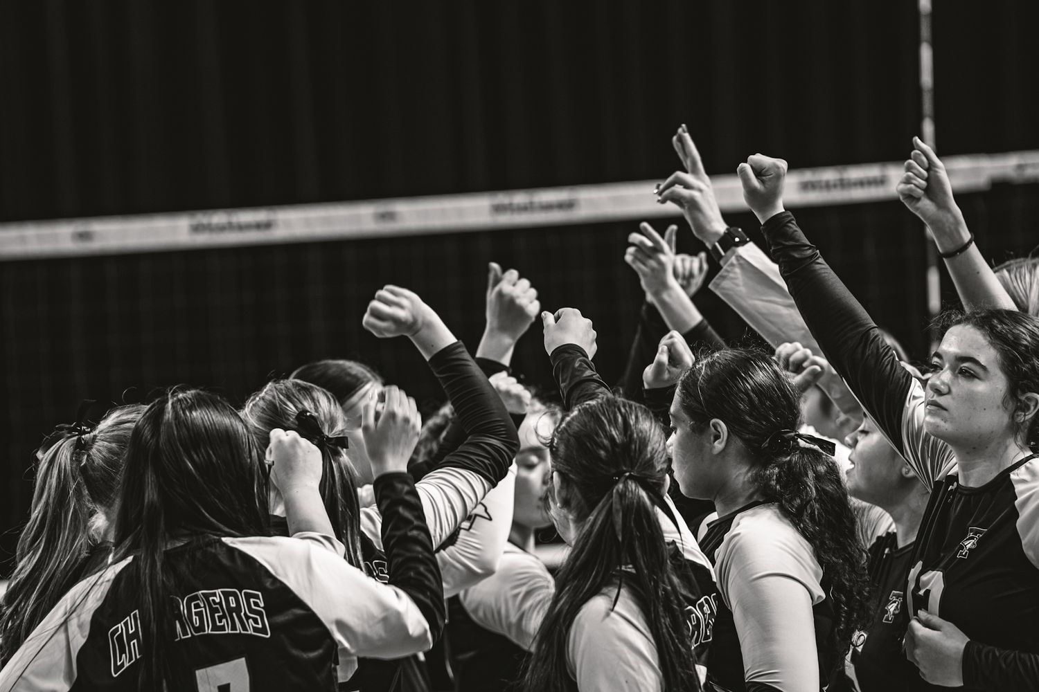 Volleyball team huddles together raising hands in black and white photo.