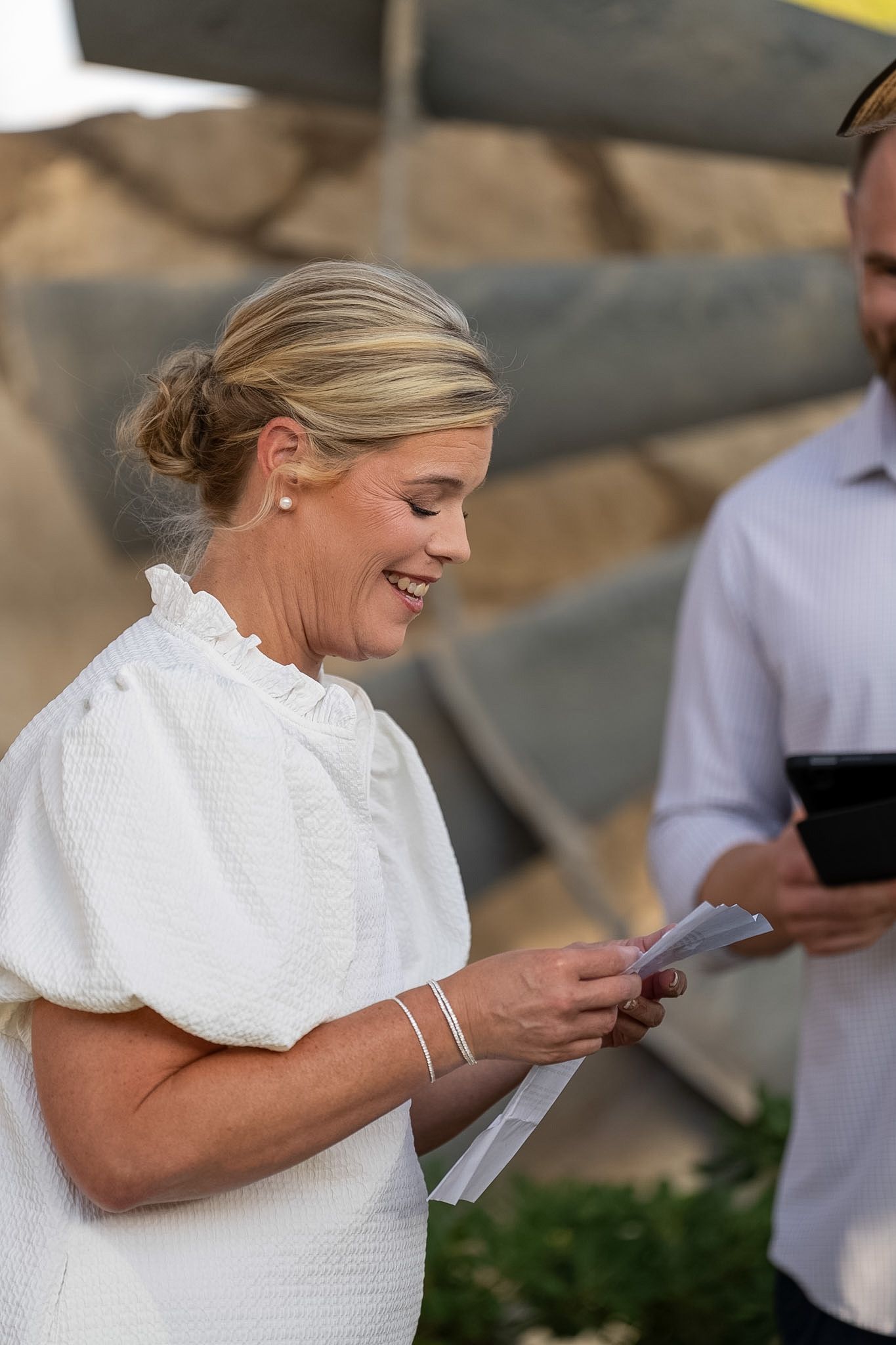 Smiling blonde woman in white dress reads a paper outdoors at a sunny event.