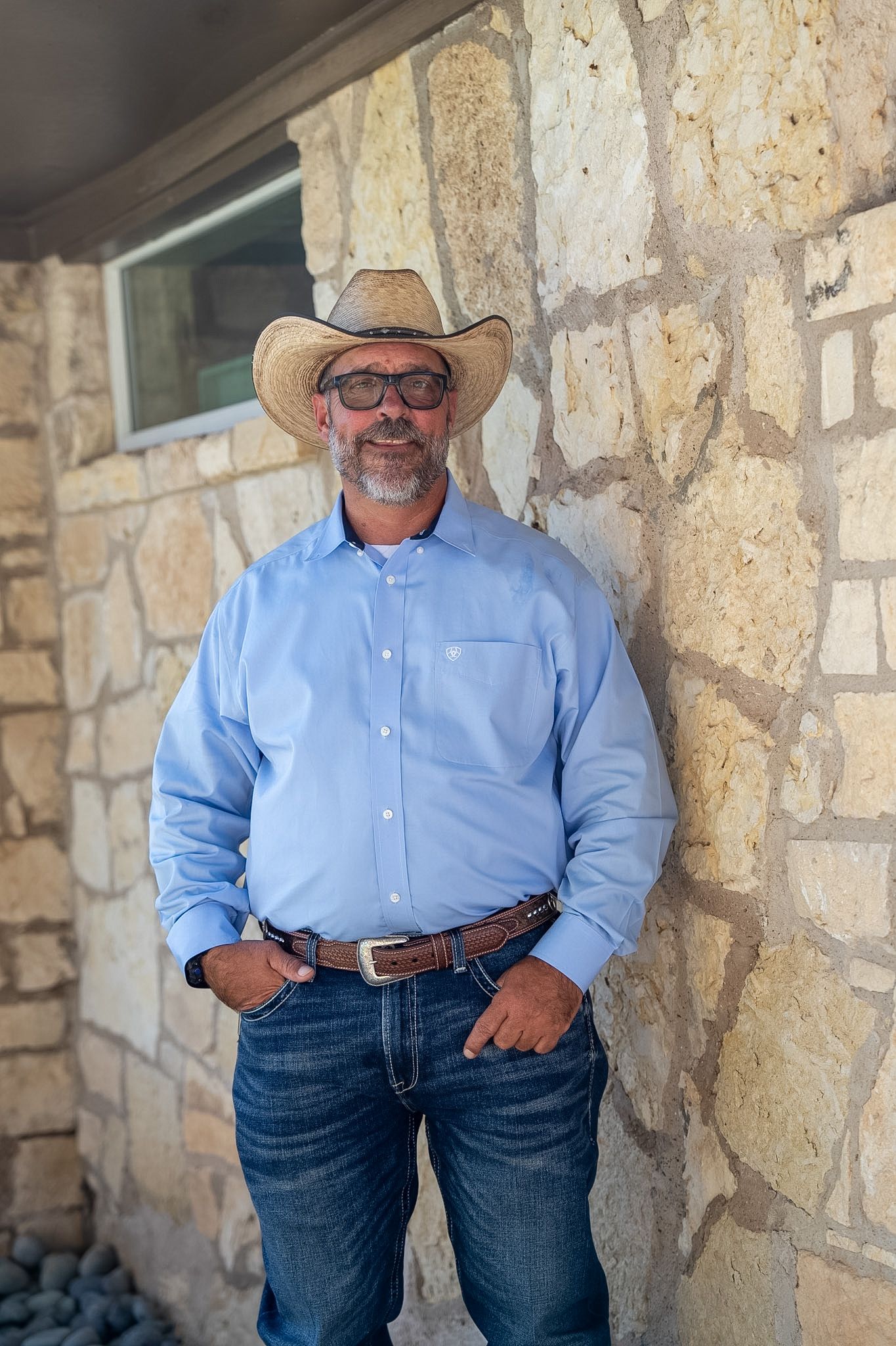 Man in cowboy hat, sunglasses, blue shirt, and jeans leaning against a stone wall outdoors.