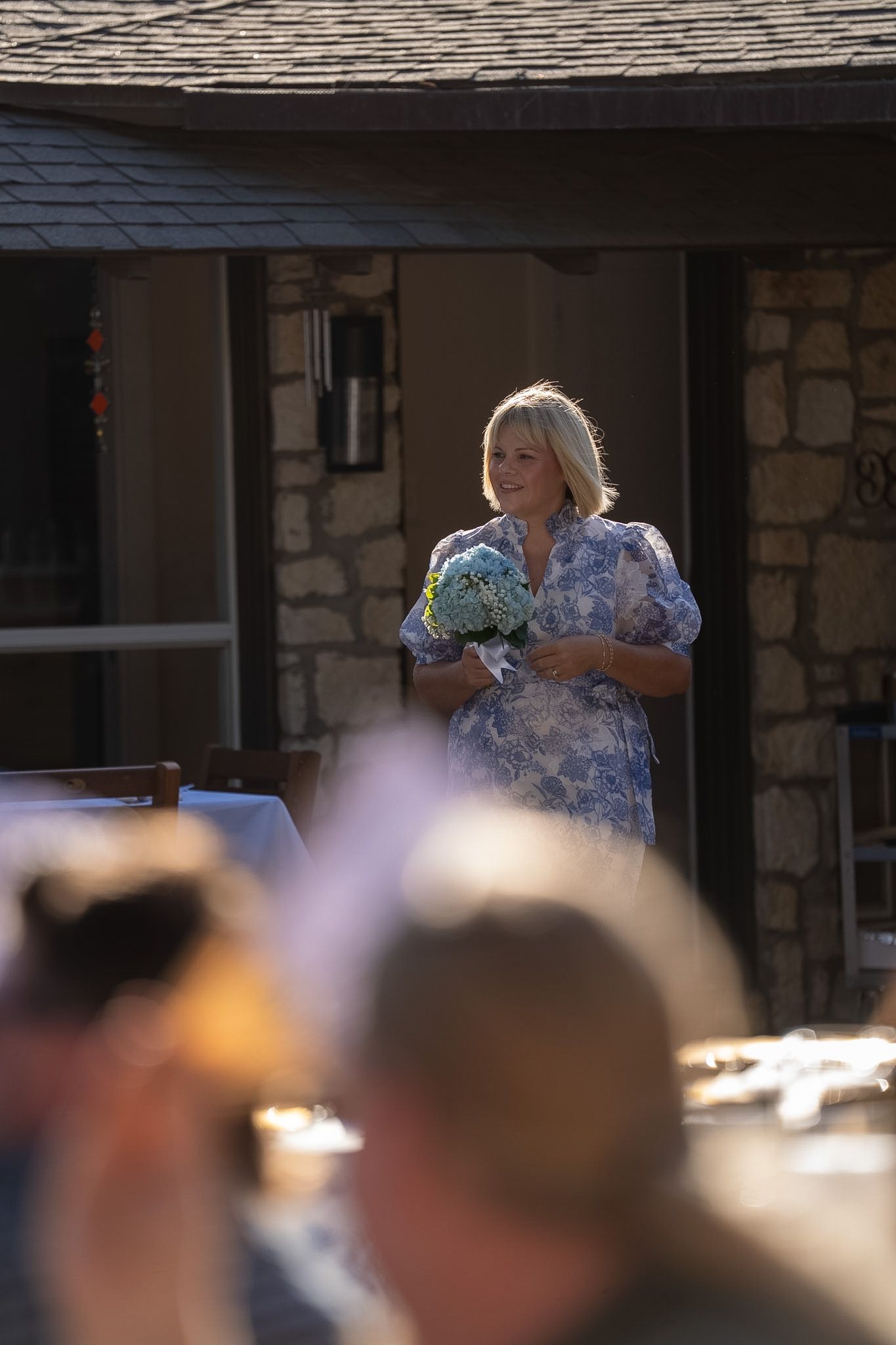 Smiling blonde woman in floral blue dress holding bouquet at outdoor wedding ceremony.