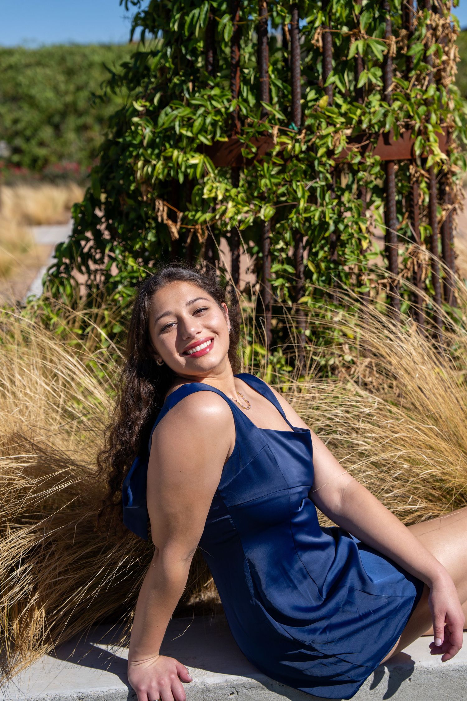 Young woman in blue dress sitting outdoors, smiling, with ornamental grasses and green ivy backdrop.