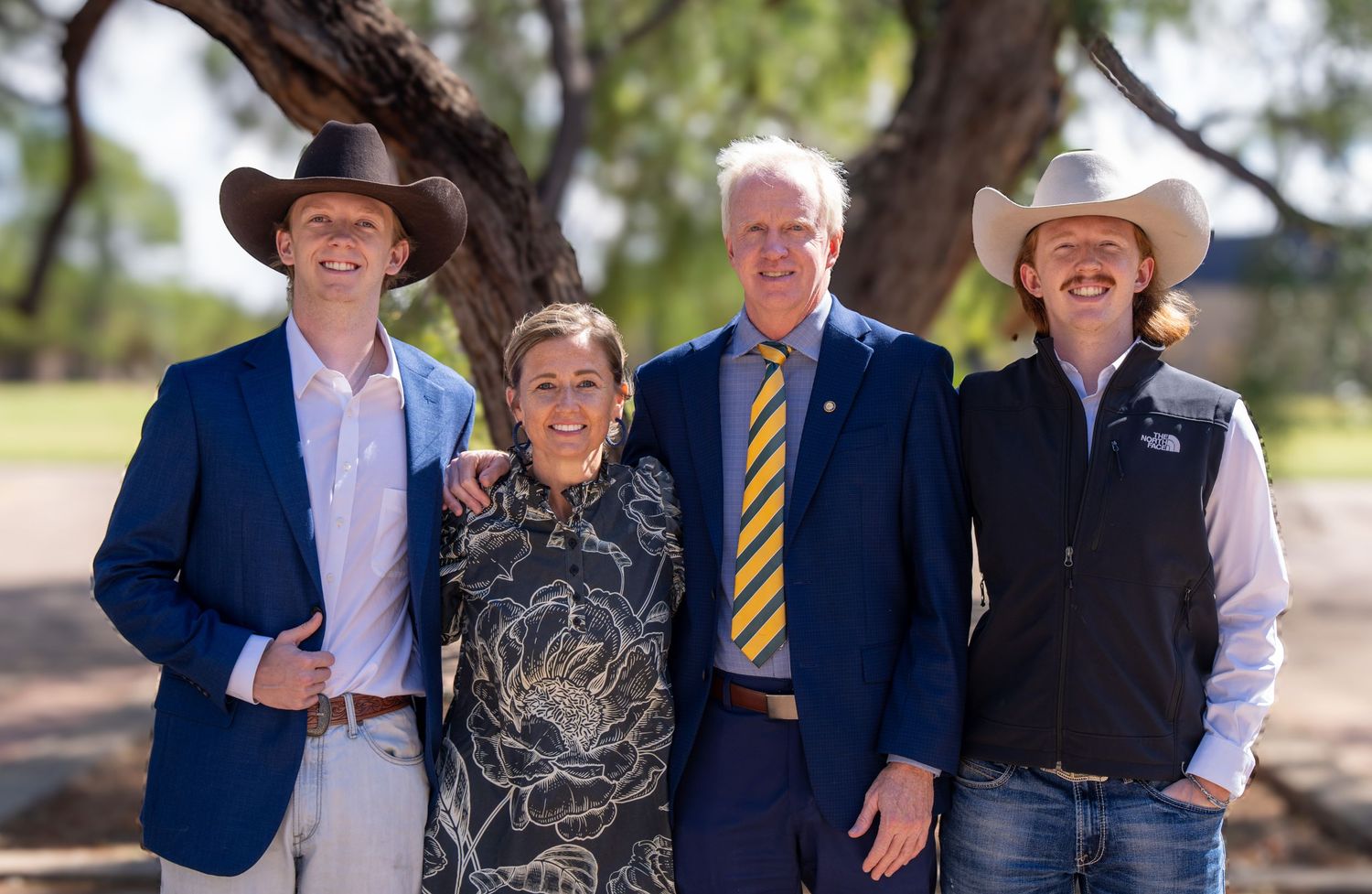 Four people smiling outdoors under a tree, two wearing cowboy hats, dressed in smart casual attire.
