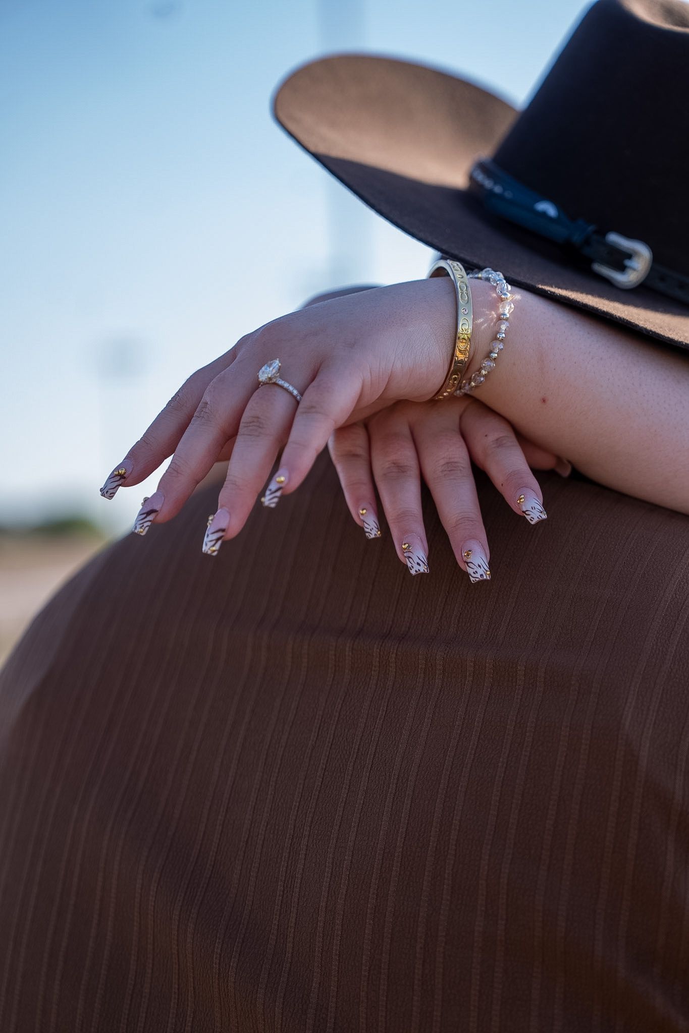 Womans hand with marquise engagement ring and gold bracelets resting on mans shoulder outdoors.