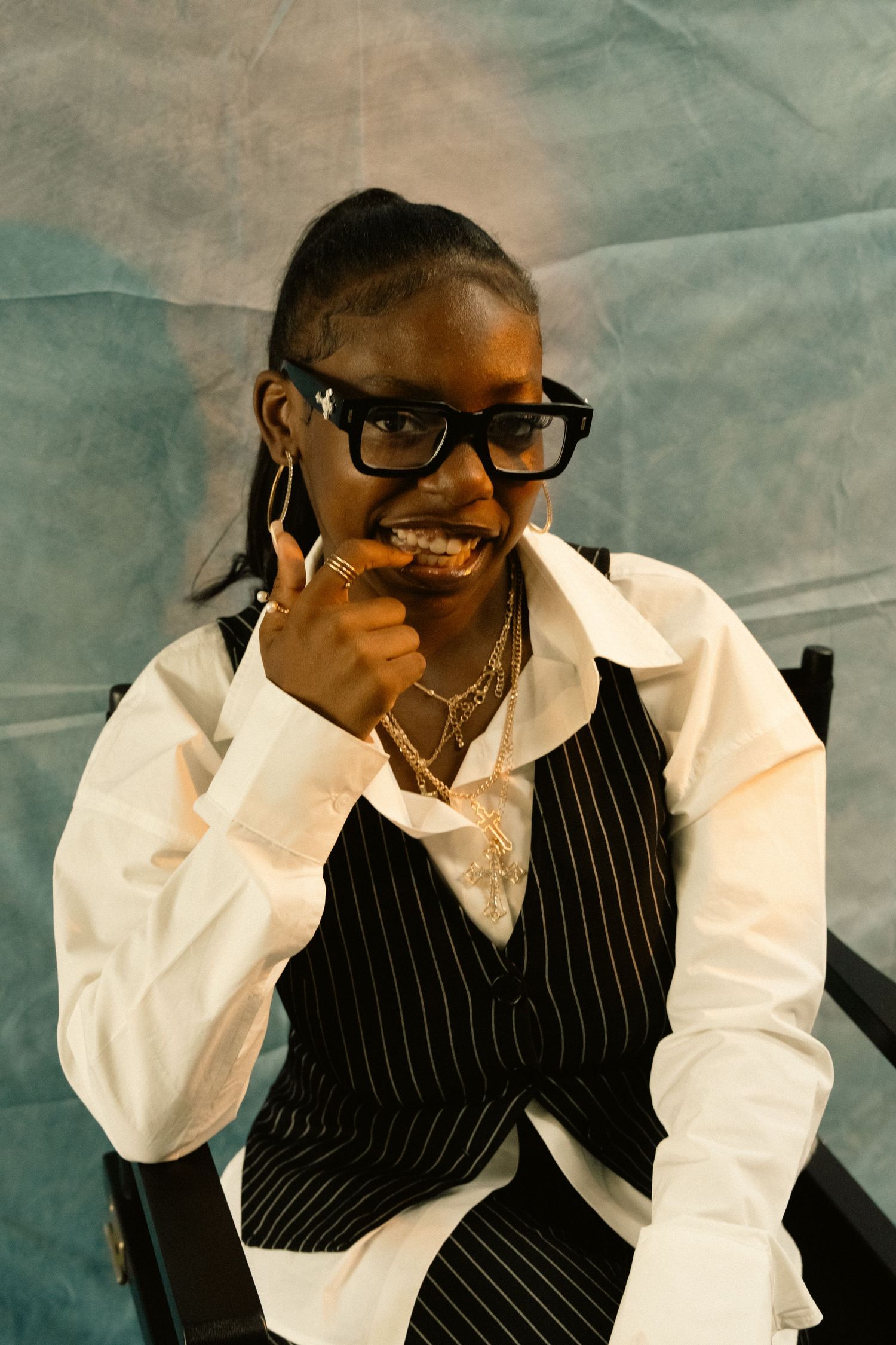 Young Black woman in pinstripe vest laughs and points finger at camera against blue backdrop.