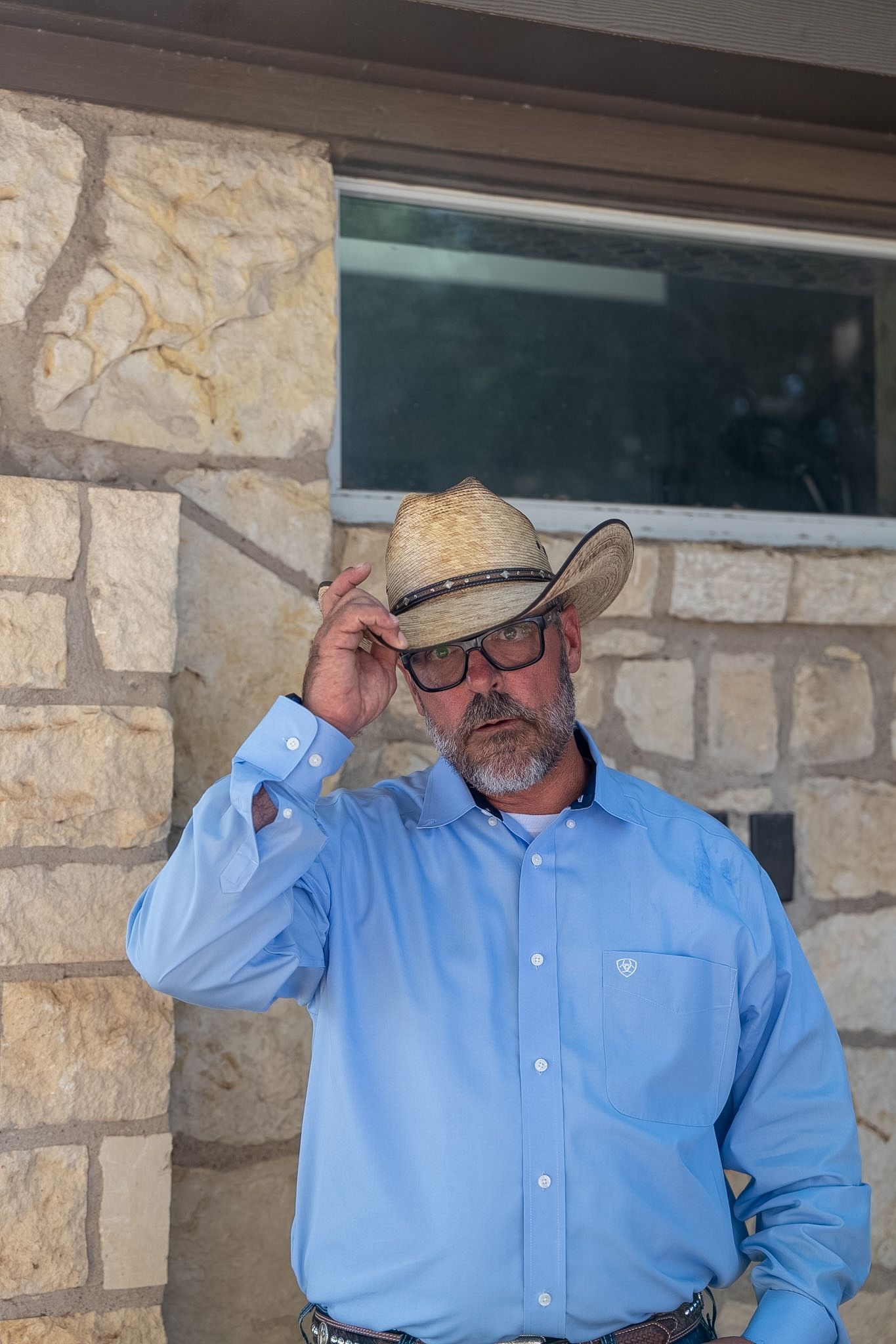 Middle-aged man in straw cowboy hat, glasses, and blue Western shirt standing near stone wall.