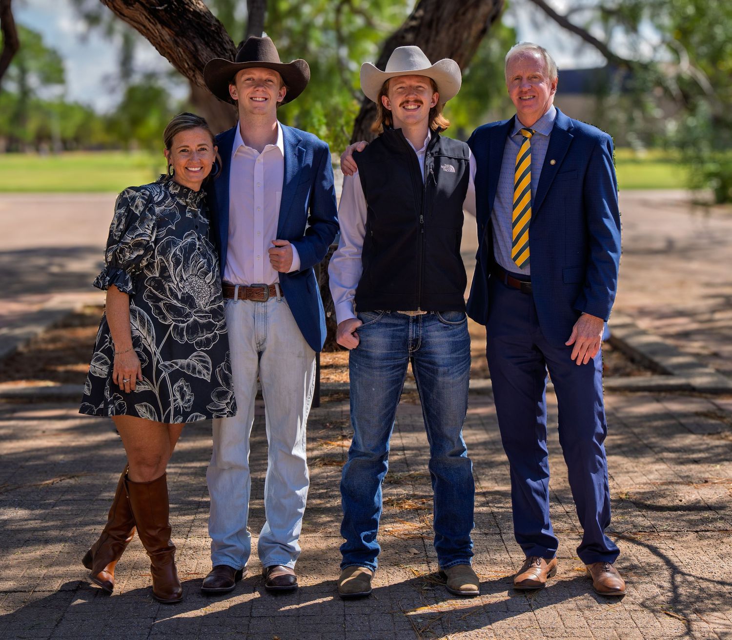 Four people posing outdoors under a tree, two wearing cowboy hats, one in a blue suit with yellow tie.