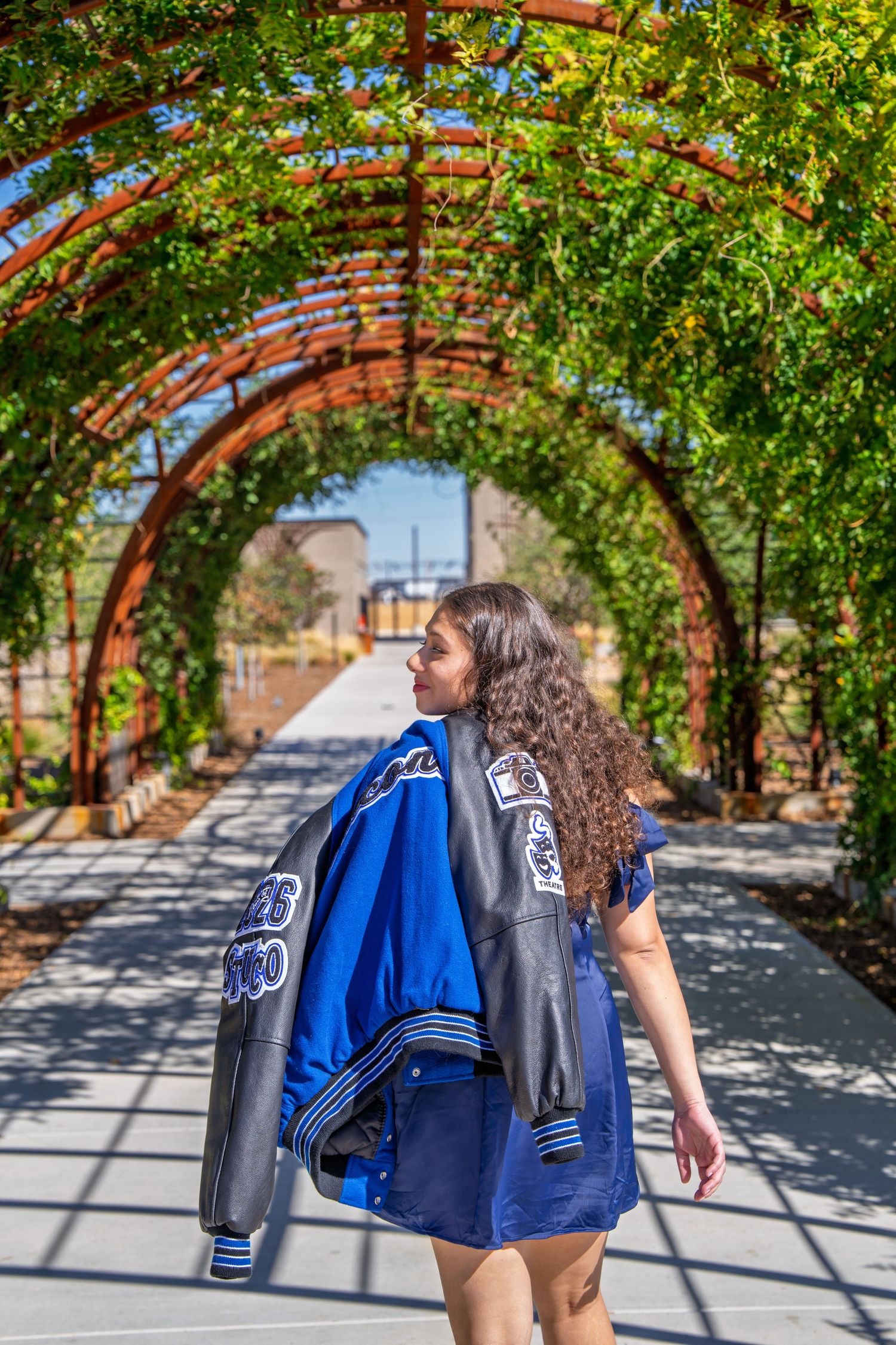 Young woman with curly hair carrying a blue varsity jacket walks under a red arched pergola with lush green vines.