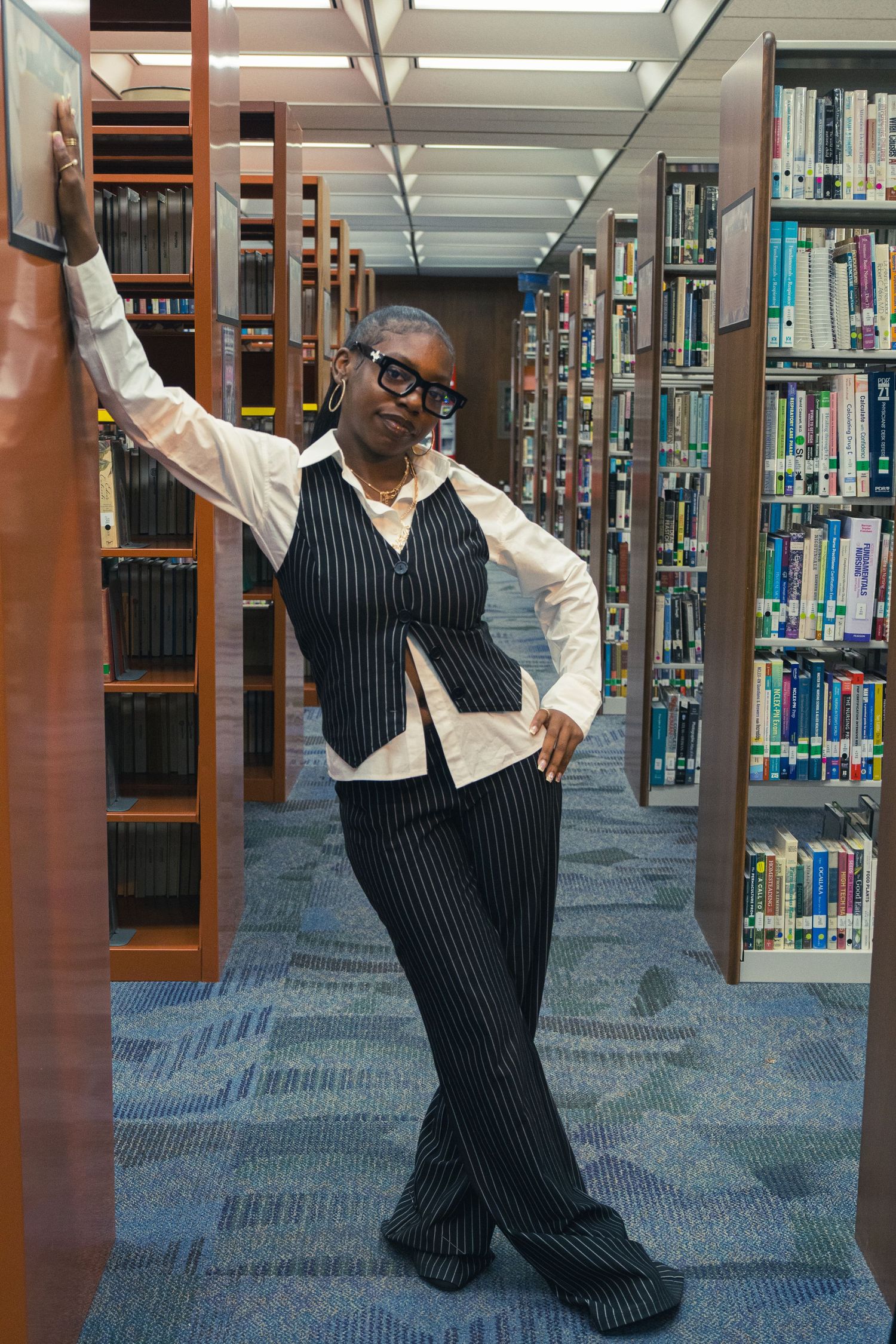 A confident Black woman in a pinstripe suit poses between library bookshelves.