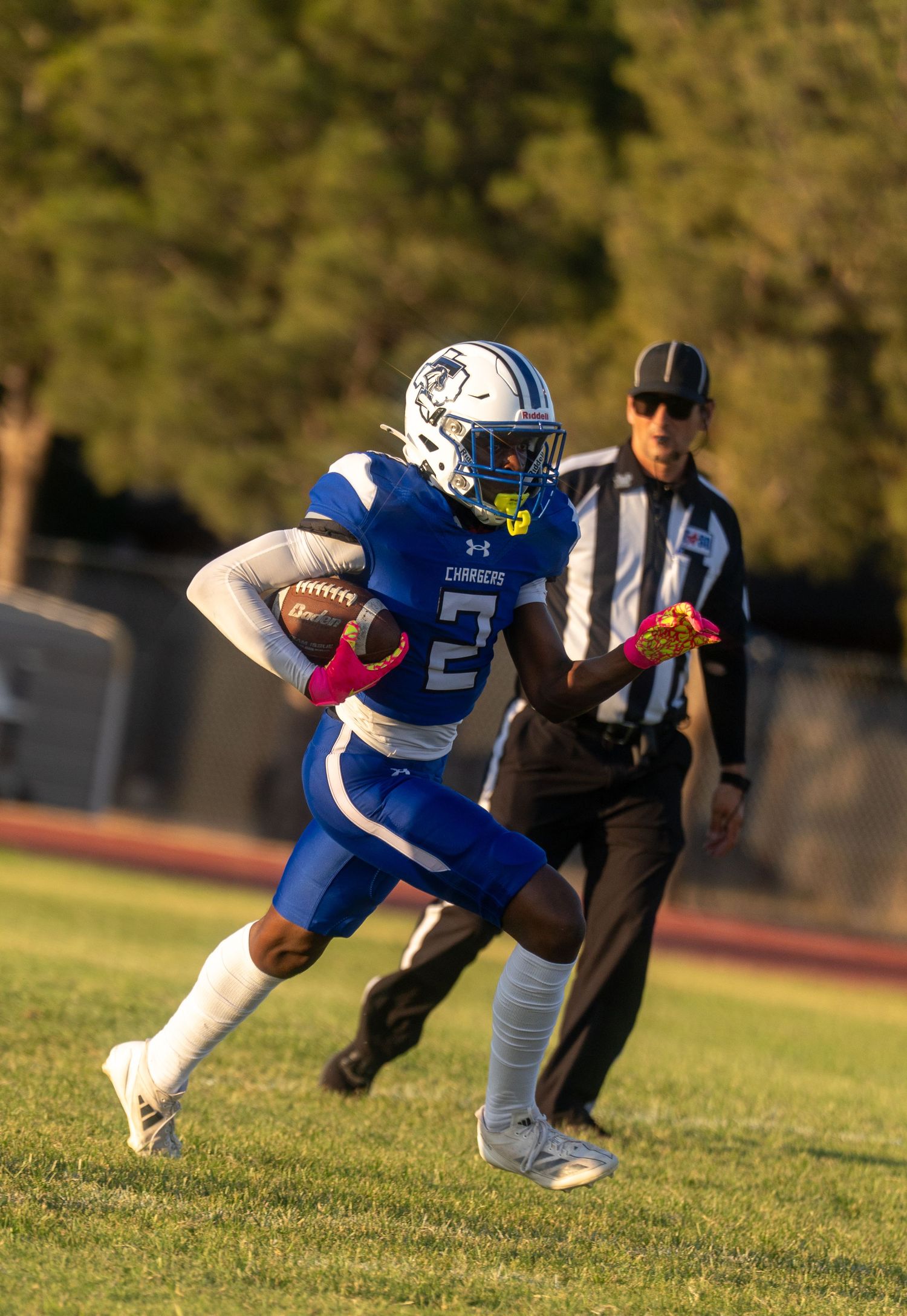 Football player #2 in blue Chargers uniform runs with the ball as a referee watches during a night game.