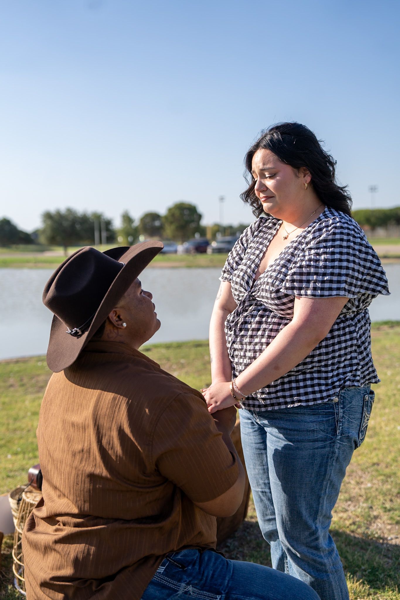 Person in cowboy hat kneels proposing to woman near lakeside on sunny day.
