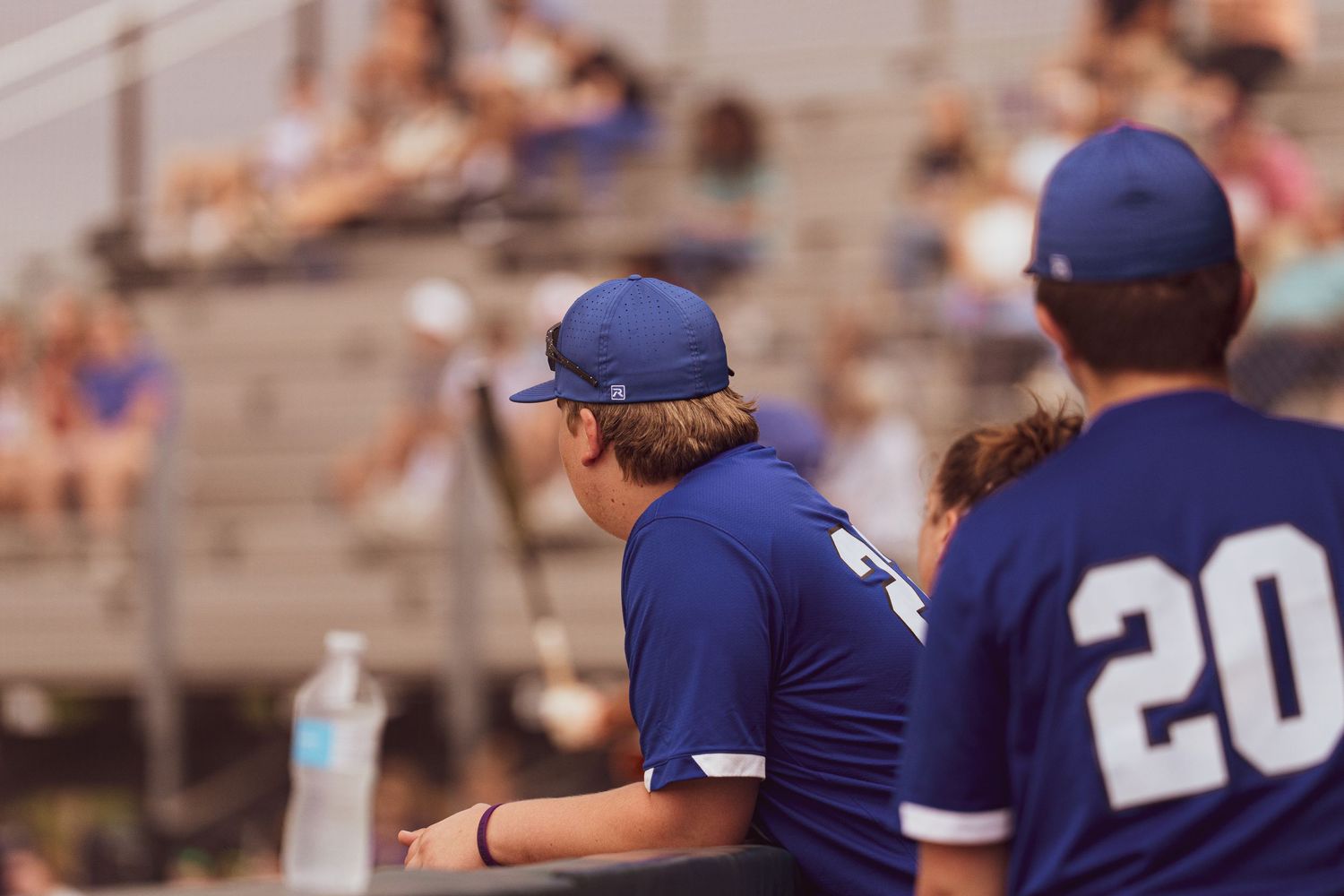 Two baseball players in blue jerseys watching game from dugout.