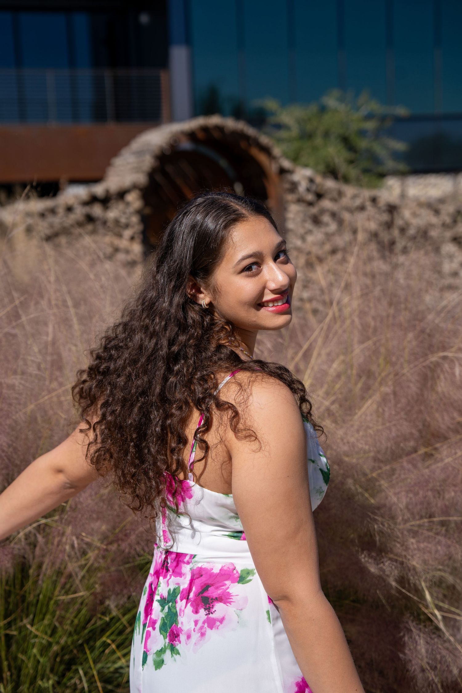 Young Latina woman with long curly hair in white floral dress smiling over shoulder outdoors.