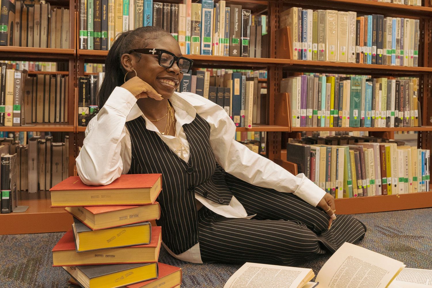 A thoughtful woman in glasses and pinstripe vest sits among stacked books in a library.