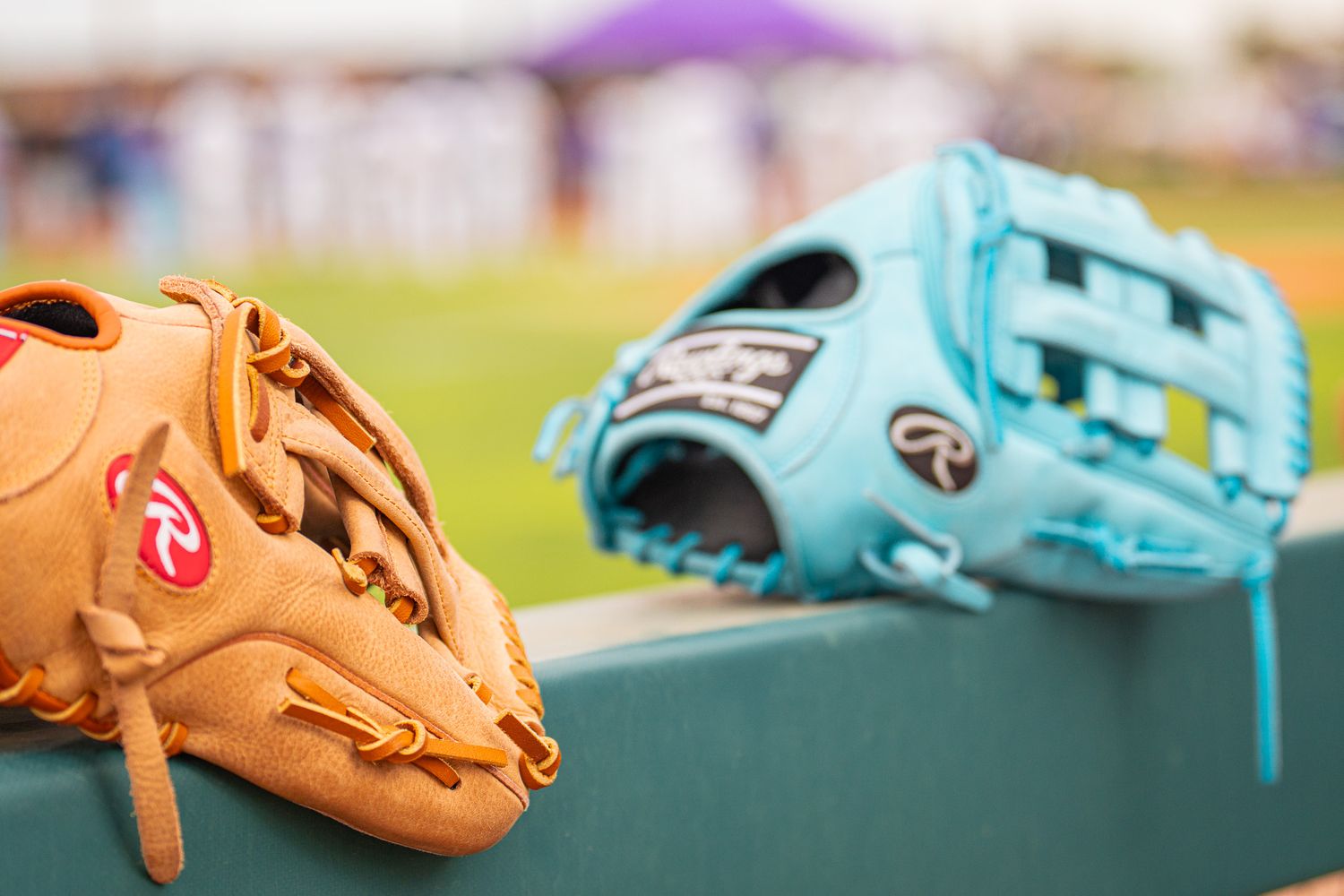 Two Rawlings baseball gloves, tan and teal, rest on a dugout ledge at a baseball field.