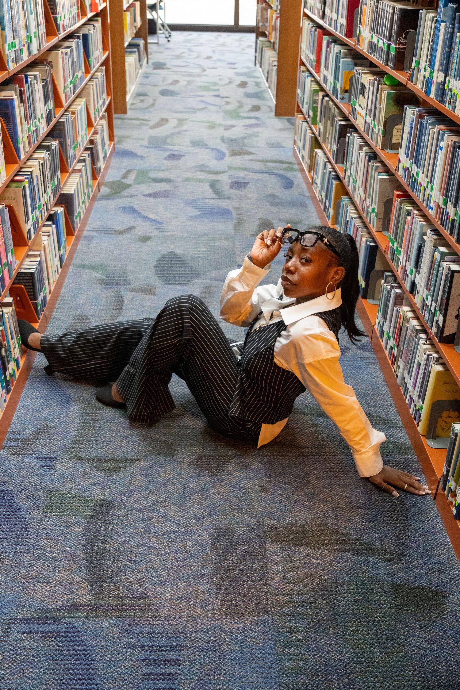 Fashionable woman in pinstripe outfit poses on library floor between bookshelves.