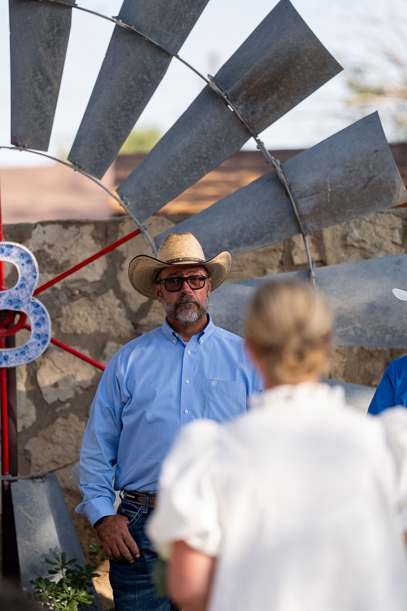 A man in a straw cowboy hat and blue shirt stands near a windmill at an outdoor ranch event.