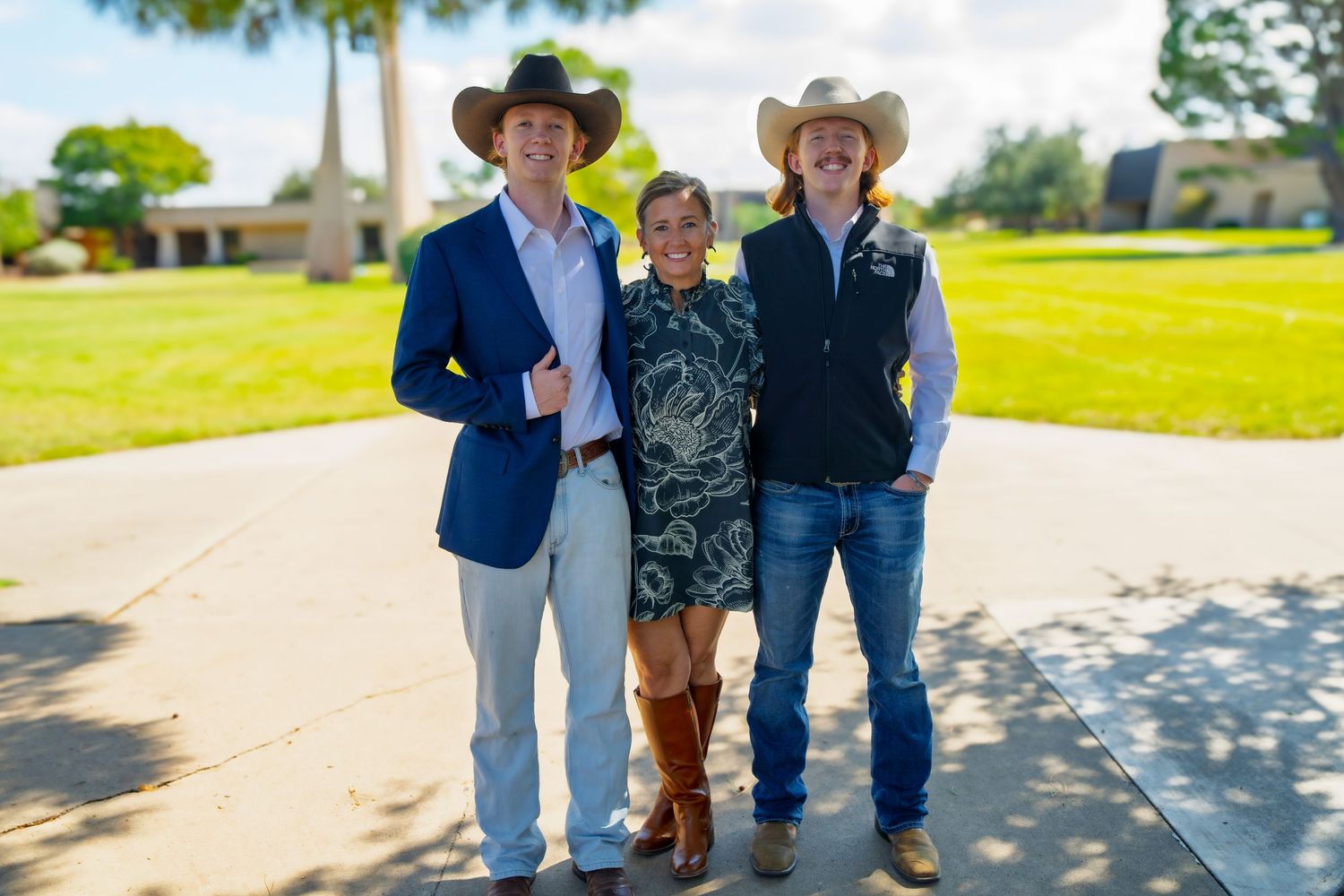 Three people in cowboy hats smiling outdoors on a sunny day near a green lawn.