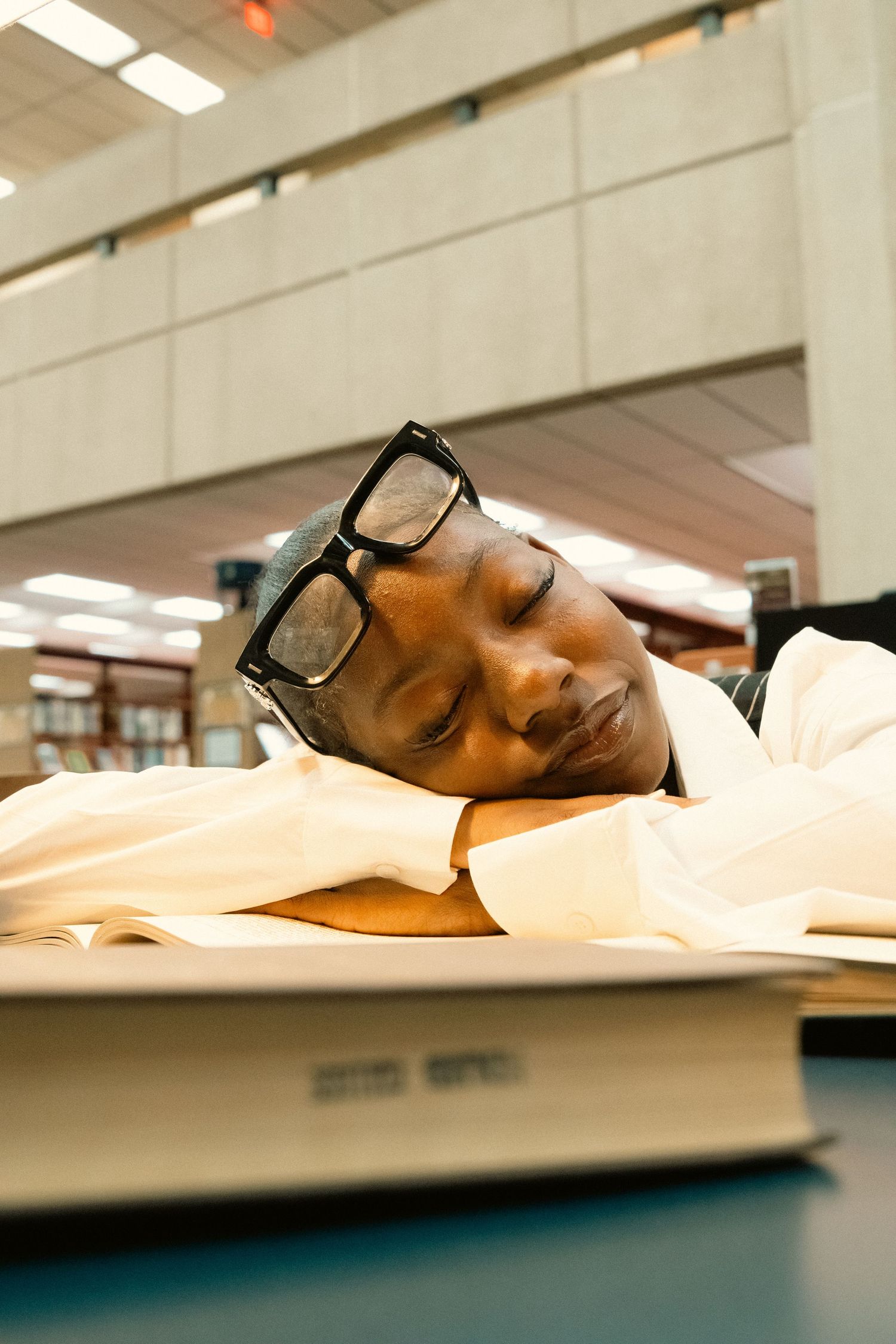 A tired student with black glasses sleeping on an open book in a library.