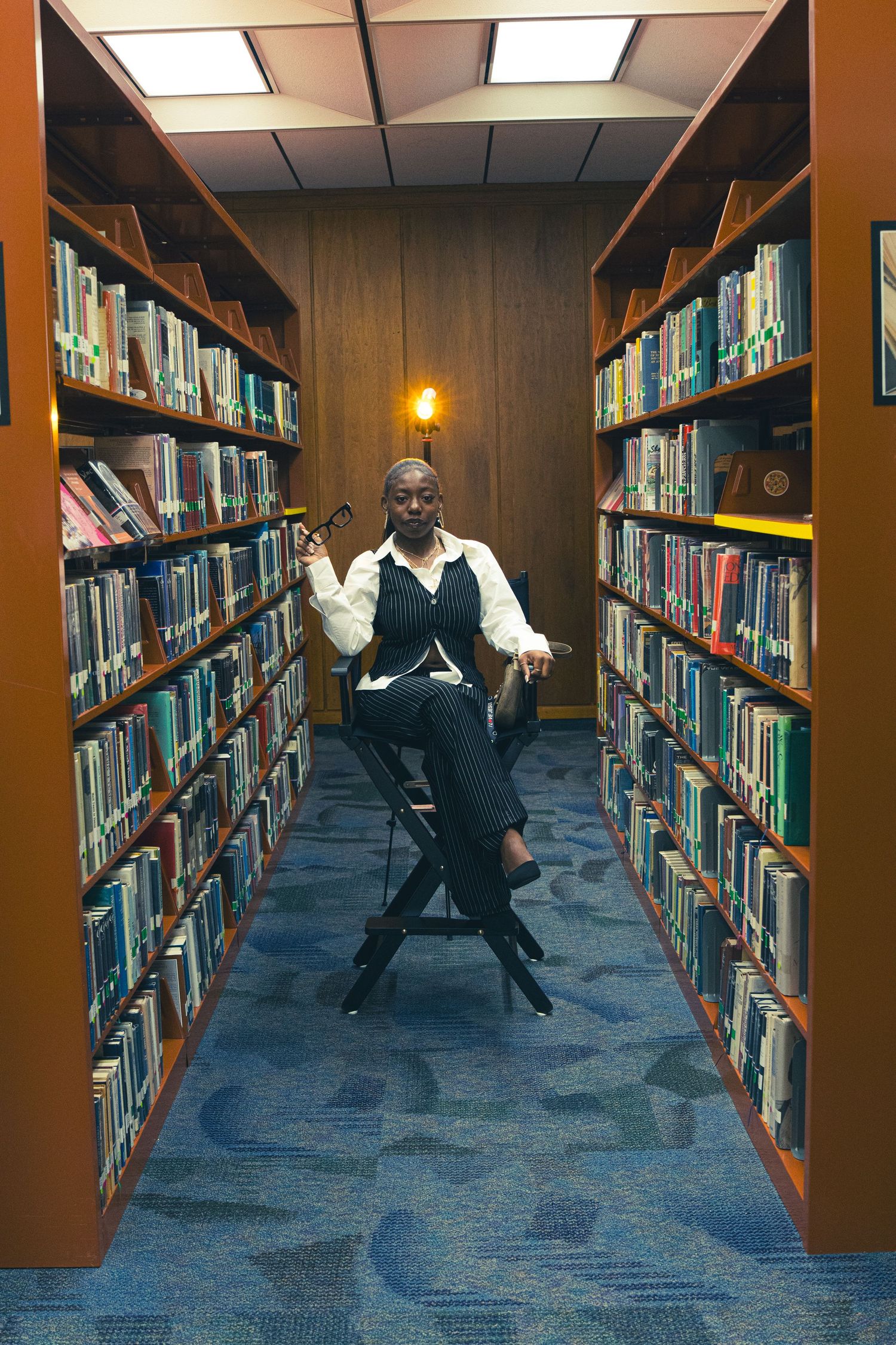 Black woman in pinstripe suit sits confidently in director chair between tall library bookshelves.