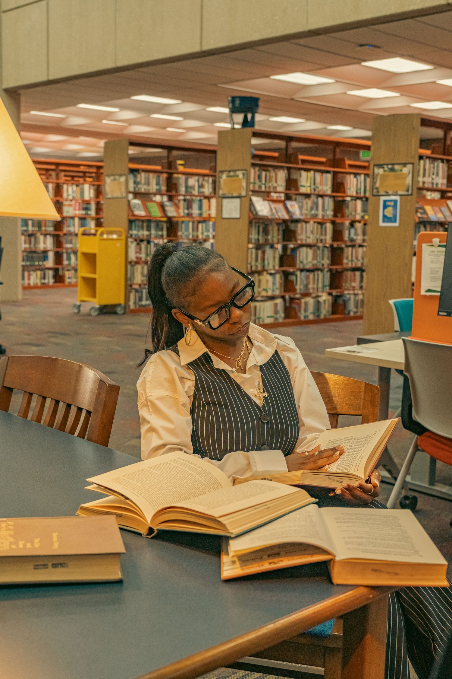 Black woman in pinstripe vest reads large open book at a library table surrounded by bookshelves.