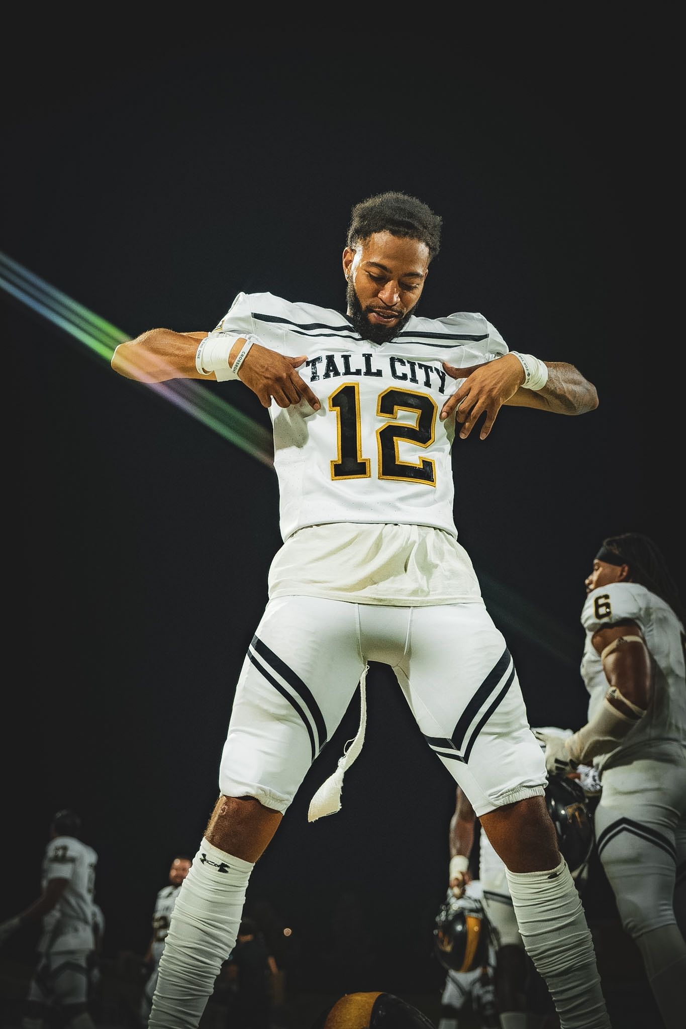 Tall City football player number 12 celebrates energetically under colorful stadium lights at night.