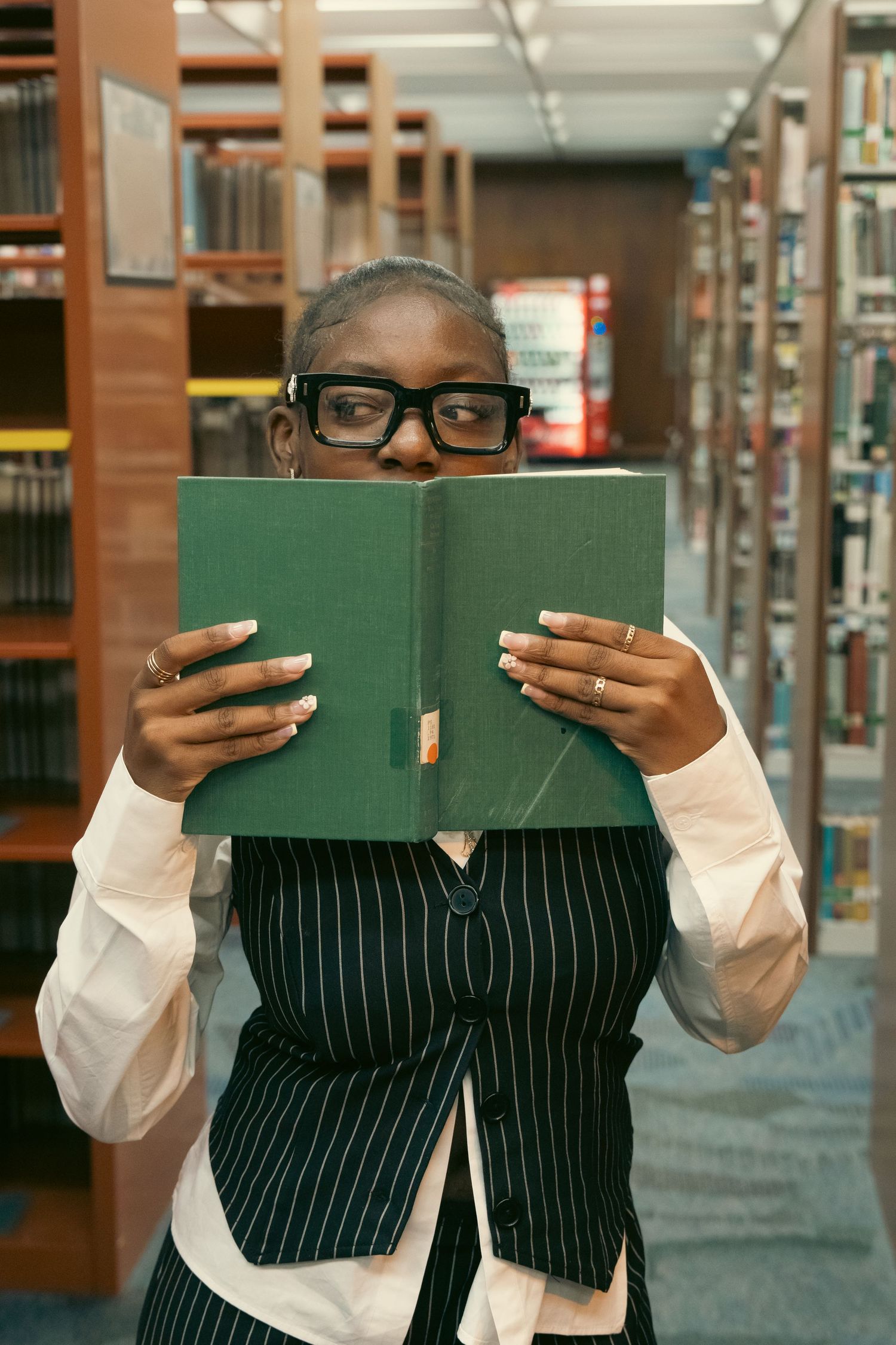 Woman in library hiding behind a green book, wearing black glasses and pinstripe vest.