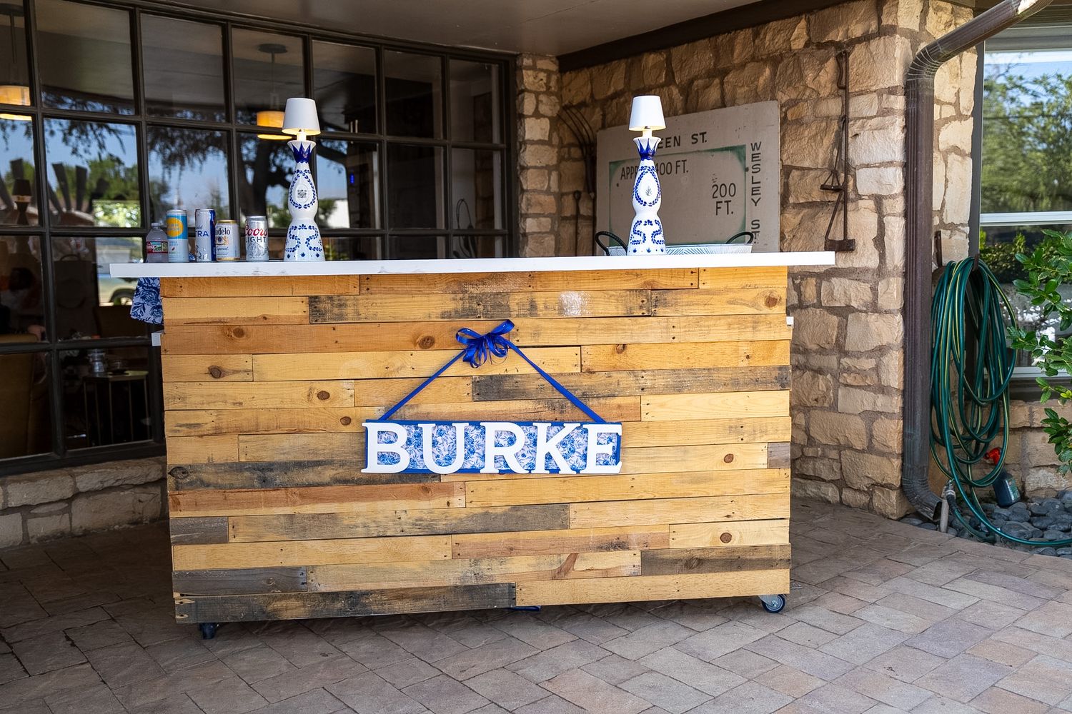 Rustic wooden pallet bar with Burke name sign, blue bow, and white lamp accents on wheels outdoors.