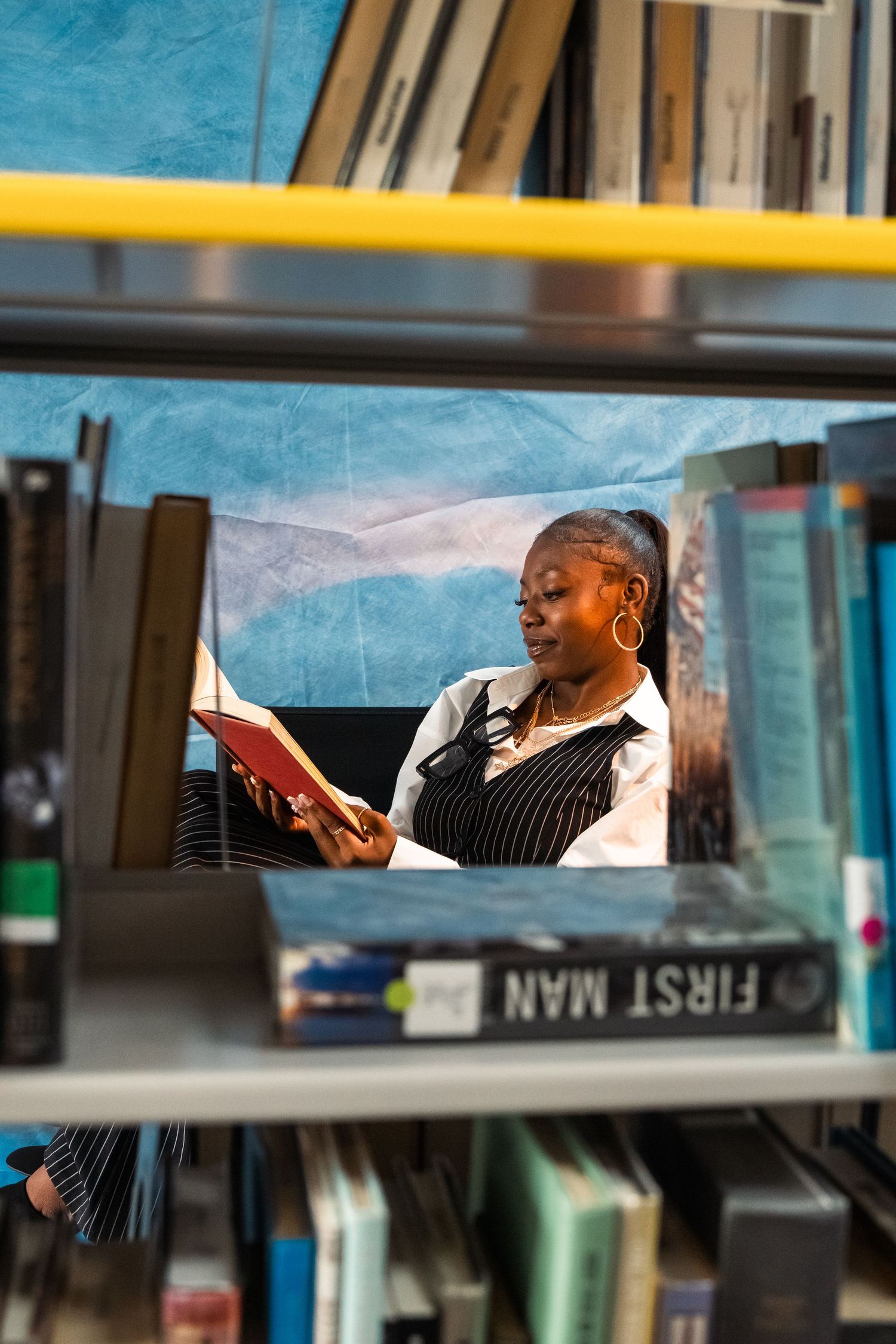 Woman reading a red book in a library, viewed through bookshelves with First Man book visible in foreground.
