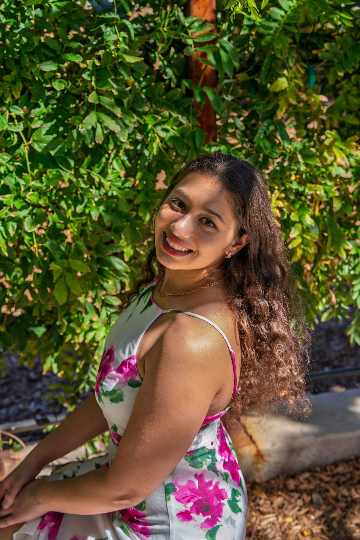 Young woman in floral dress smiling outdoors among lush green foliage on a sunny day.