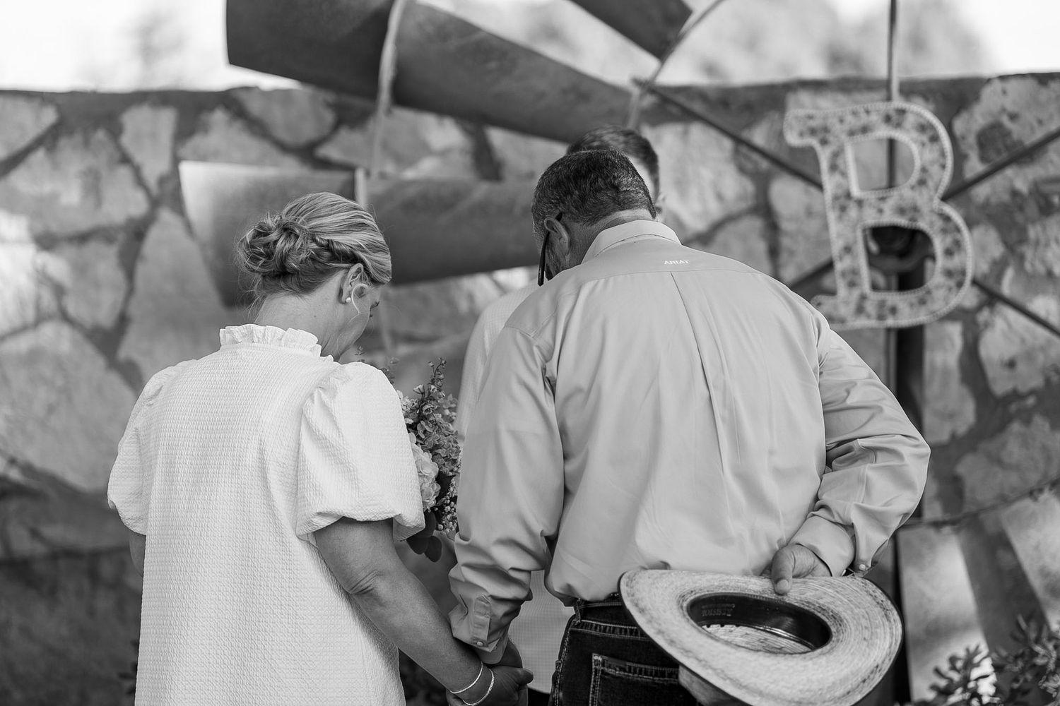 Black and white photo of two people holding hands near a decorative stone wall with a metal letter B.