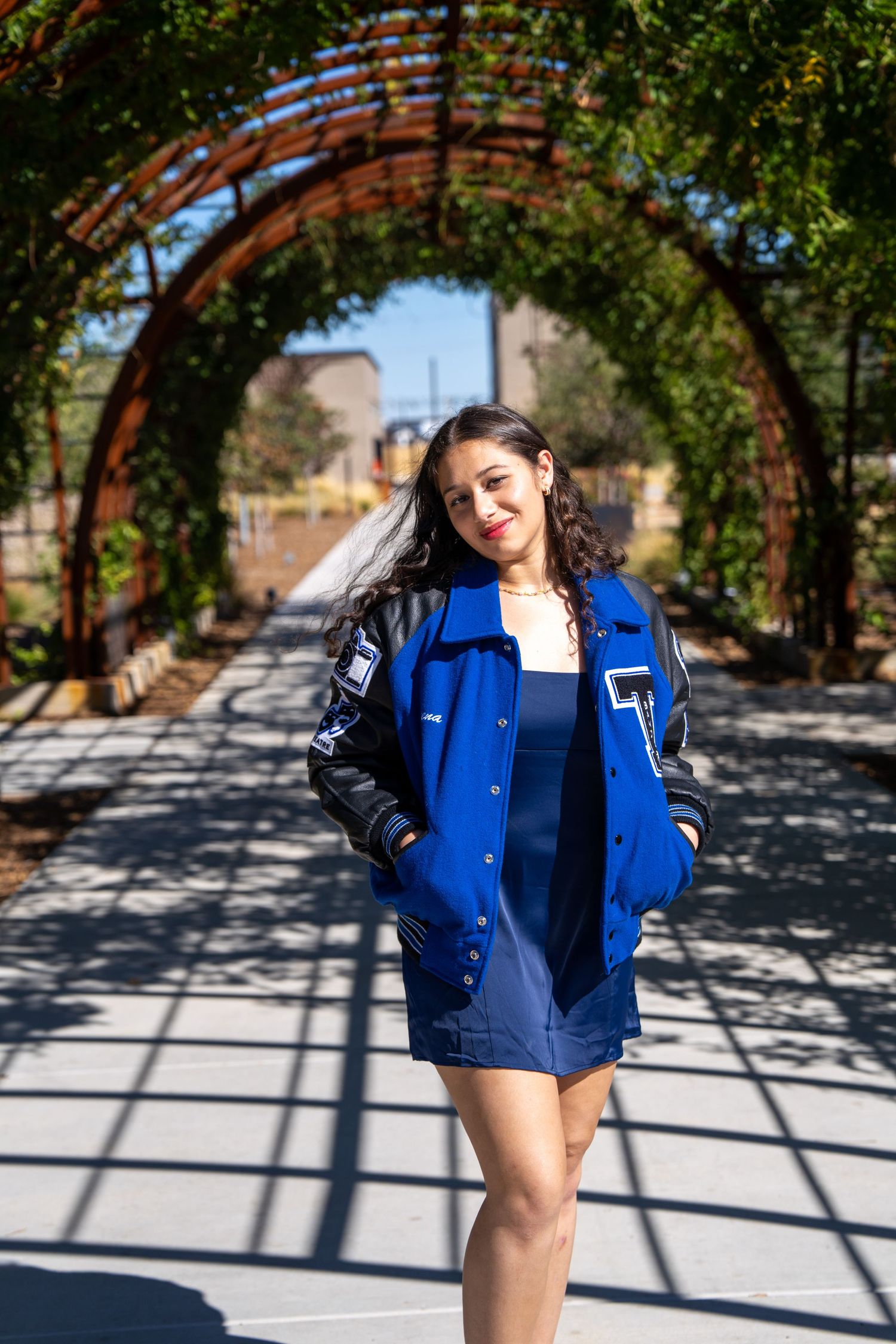 Young woman in blue varsity jacket and mini dress poses under rustic arched trellis in sunny outdoor garden path.