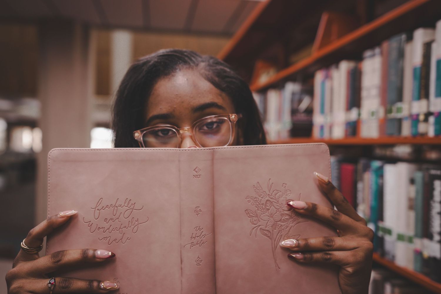 Young Black woman with glasses peering over a pink hardcover book in a library with shelves in background.
