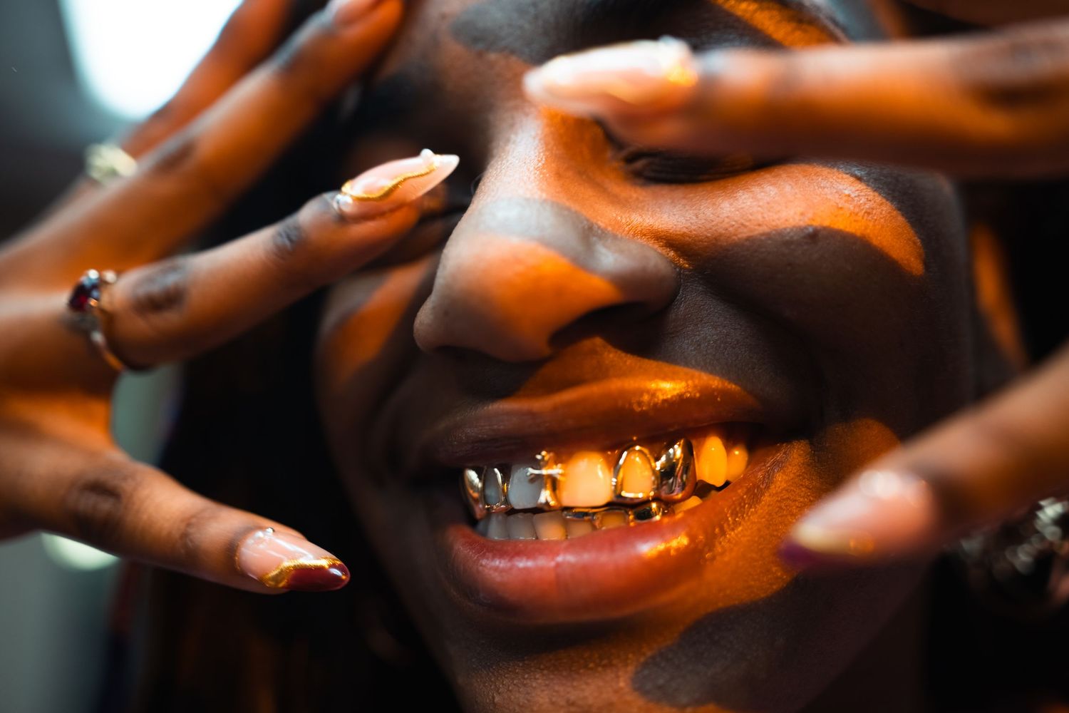 Close-up of a smiling person with gold grillz and long nails framing their face in orange light.
