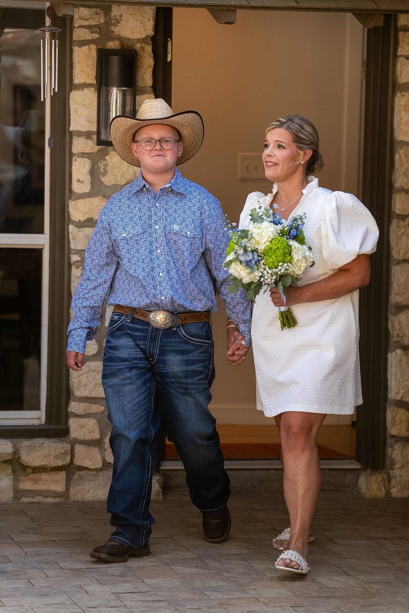 A bride in a white dress holding a blue and white bouquet walks hand-in-hand with a groom in a cowboy hat and blue shirt.