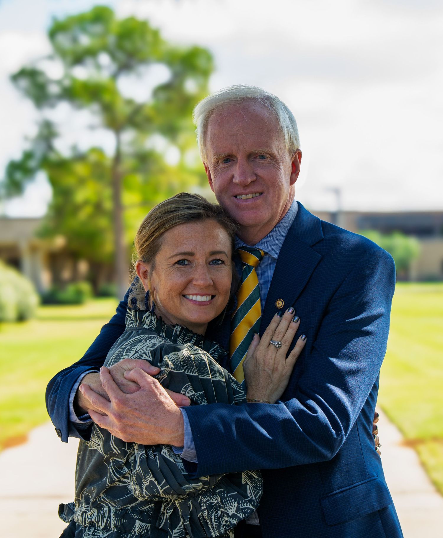 A smiling couple embraces outdoors, man in blue blazer with gold striped tie, woman in floral top, green trees behind.