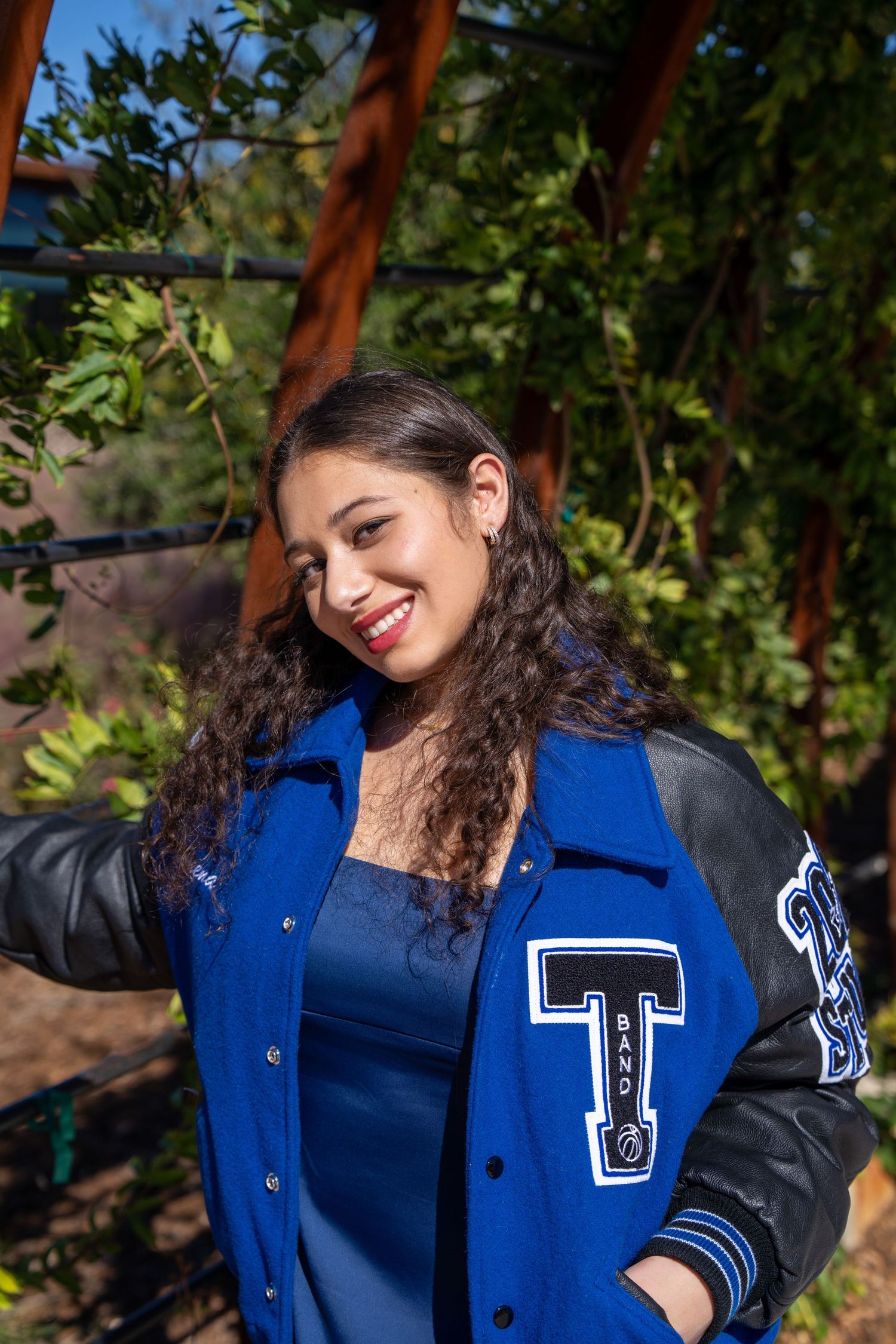 Young woman smiling in a blue varsity jacket with black leather sleeves, posing outdoors near green foliage.